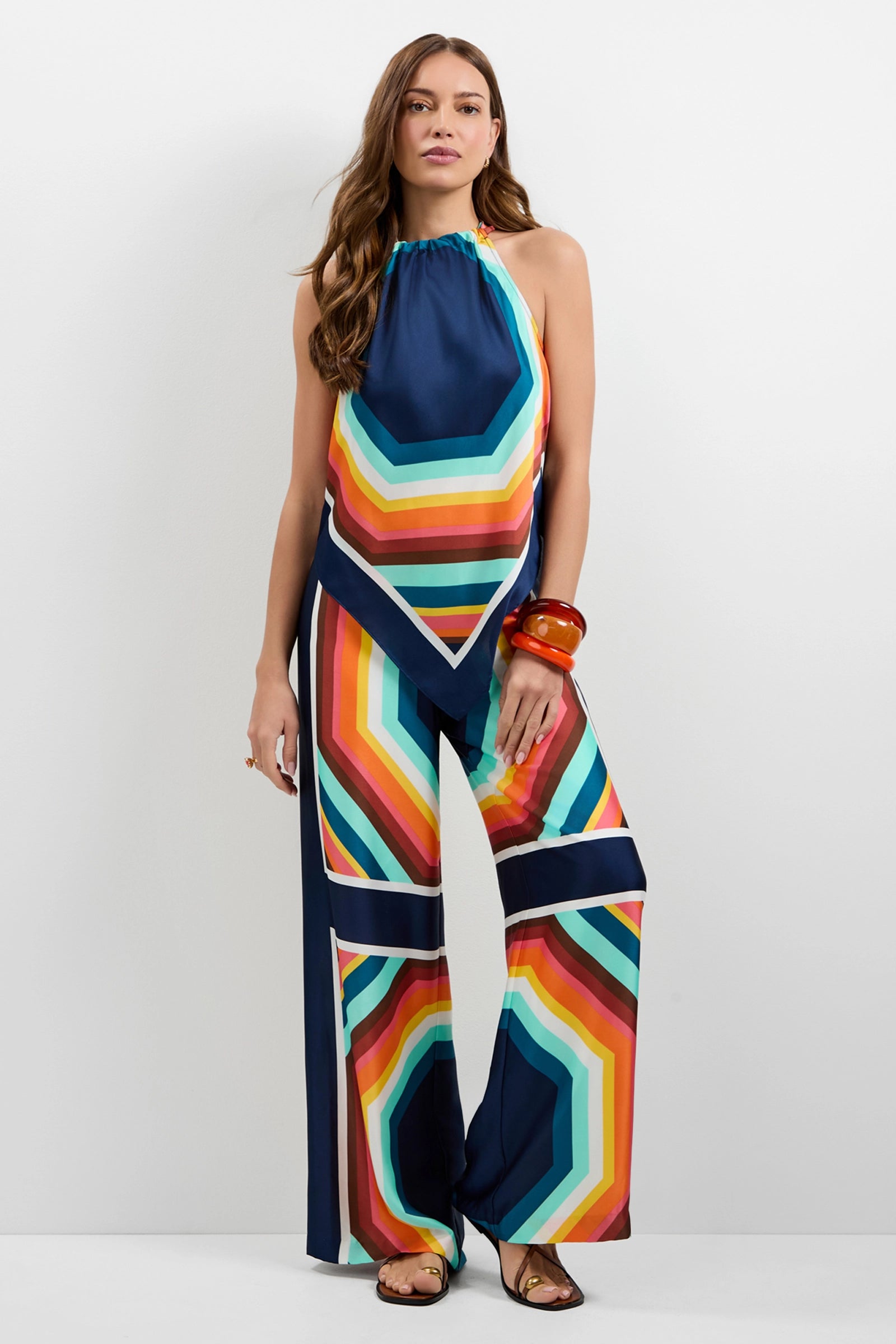 A woman stands against a plain white background, wearing Lanay Siena Wide-Leg Pants with a colorful geometric pattern, styled with loose wavy hair, large orange bangles, and sandals.