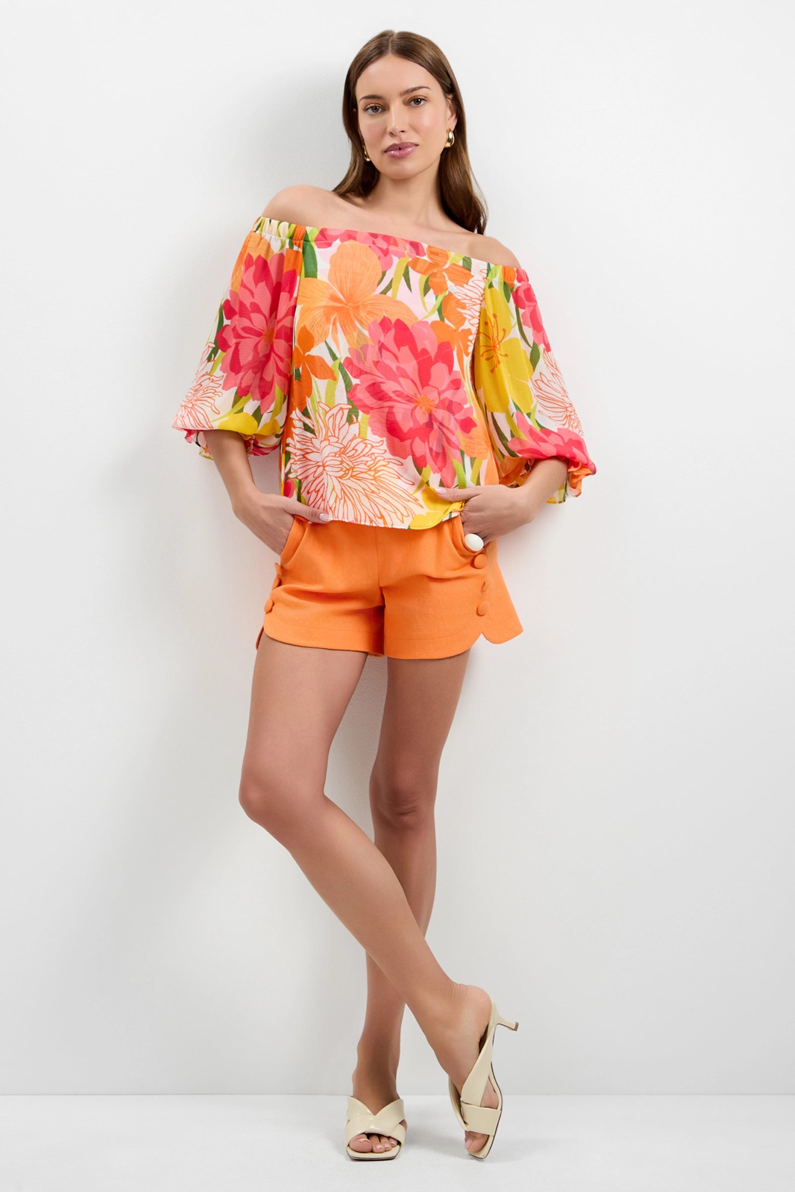 A woman with long hair poses relaxed against a white wall, wearing the Vittoria Off-Shoulder Top featuring large pink, yellow, and orange flowers, paired with bright orange shorts and beige open-toe heels.