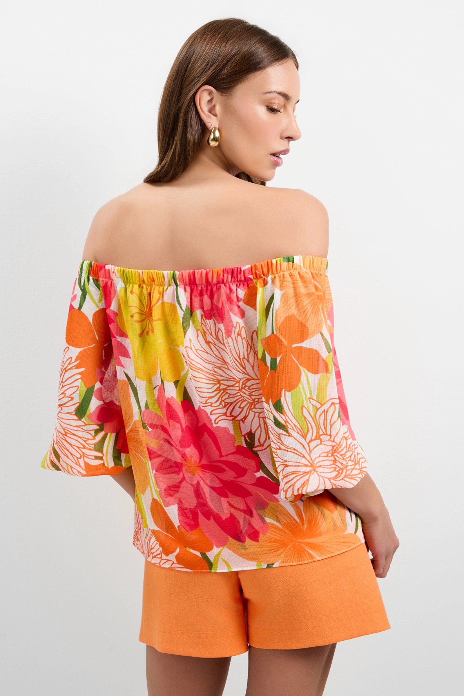 A woman with straight brown hair and gold hoop earrings stands against a white background, facing away and looking over her shoulder, wearing the colorful Vittoria Off-Shoulder Top and orange shorts.