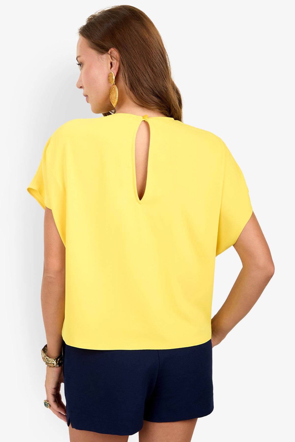 Woman wearing the Scodelle Top, a yellow short-sleeve blouse with back keyhole detail, paired with dark navy shorts, facing away from the camera. She has long brown hair, gold earrings, and a bracelet.