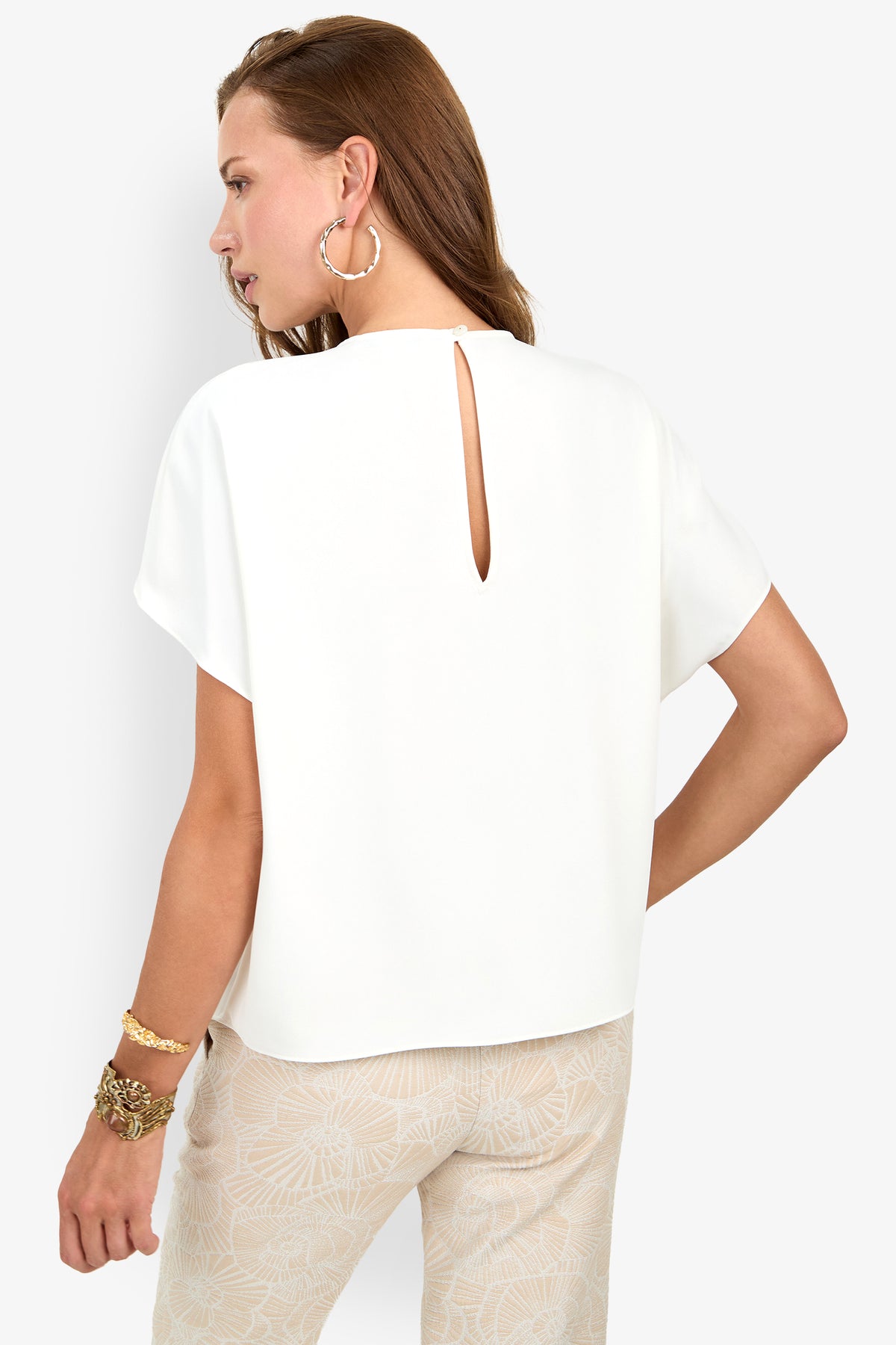 Woman facing away, wearing the Scodelle Top—a white short-sleeve blouse with a back keyhole detail—and patterned beige pants. She has hoop earrings, gold bracelets, and long brown hair tucked behind one ear.