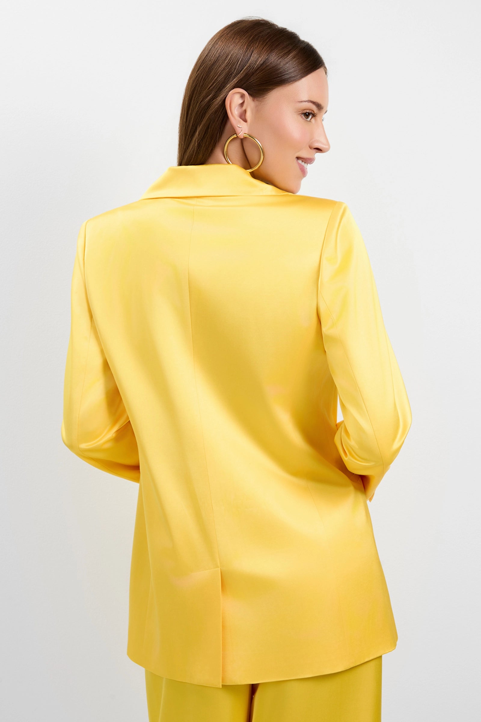 A woman with straight brown hair, large hoop earrings, and a bright yellow Giardino Satin Blazer with matching pants stands facing away, looking over her shoulder and smiling.