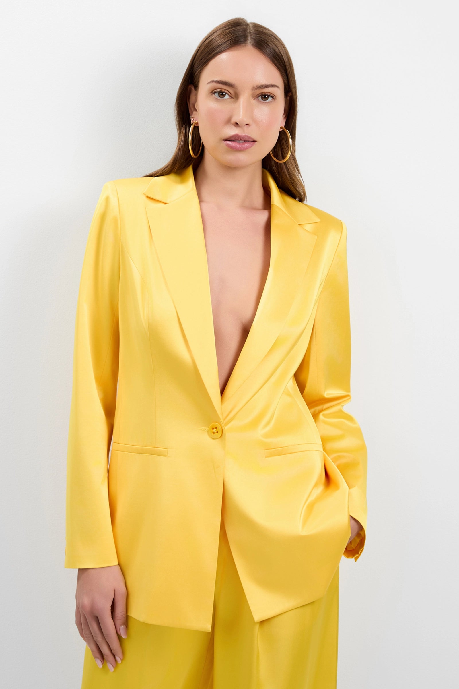 A woman with long brown hair wears the Giardino Satin Blazer, a bright yellow suit with a deep neckline. She poses against a plain white background, one hand in her pocket, accessorized with hoop earrings.