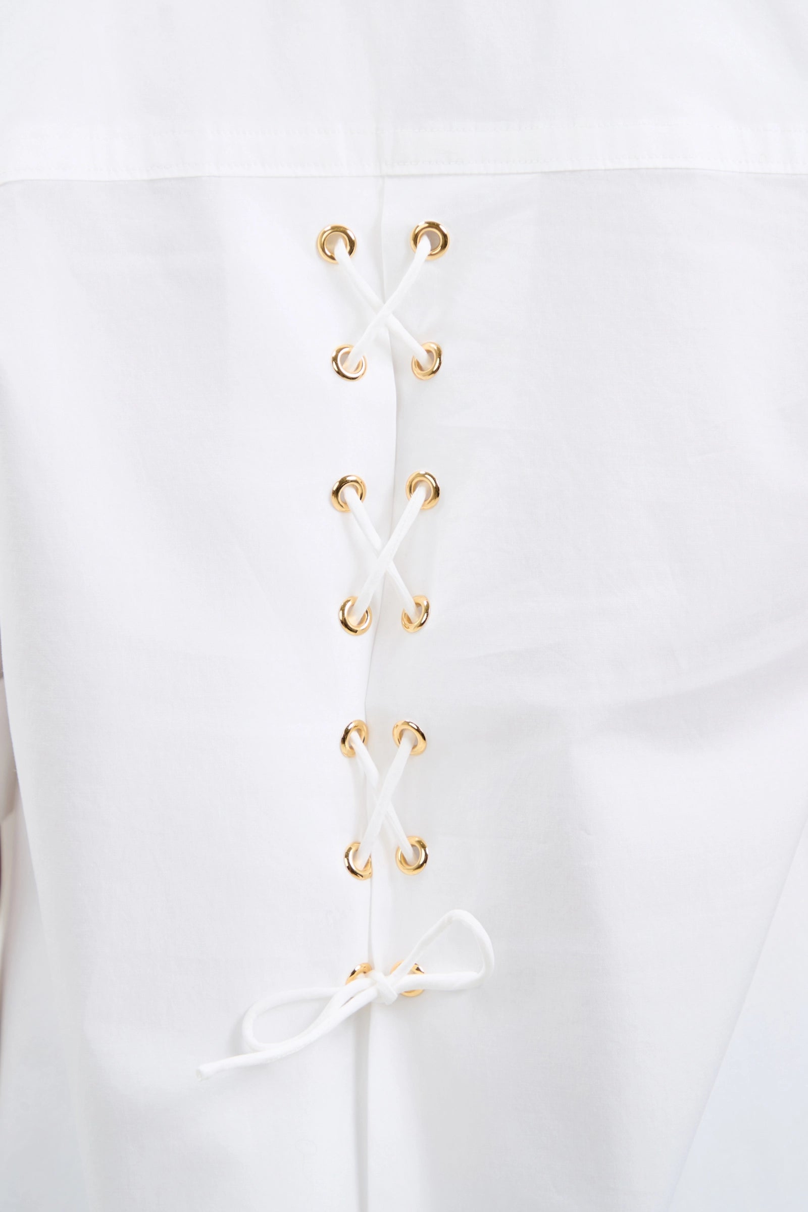 Close-up of the Triennale Lace-Up Back Top: white fabric with gold metal eyelets and white laces in a crisscross corset-inspired pattern, tied in a bow at the bottom.