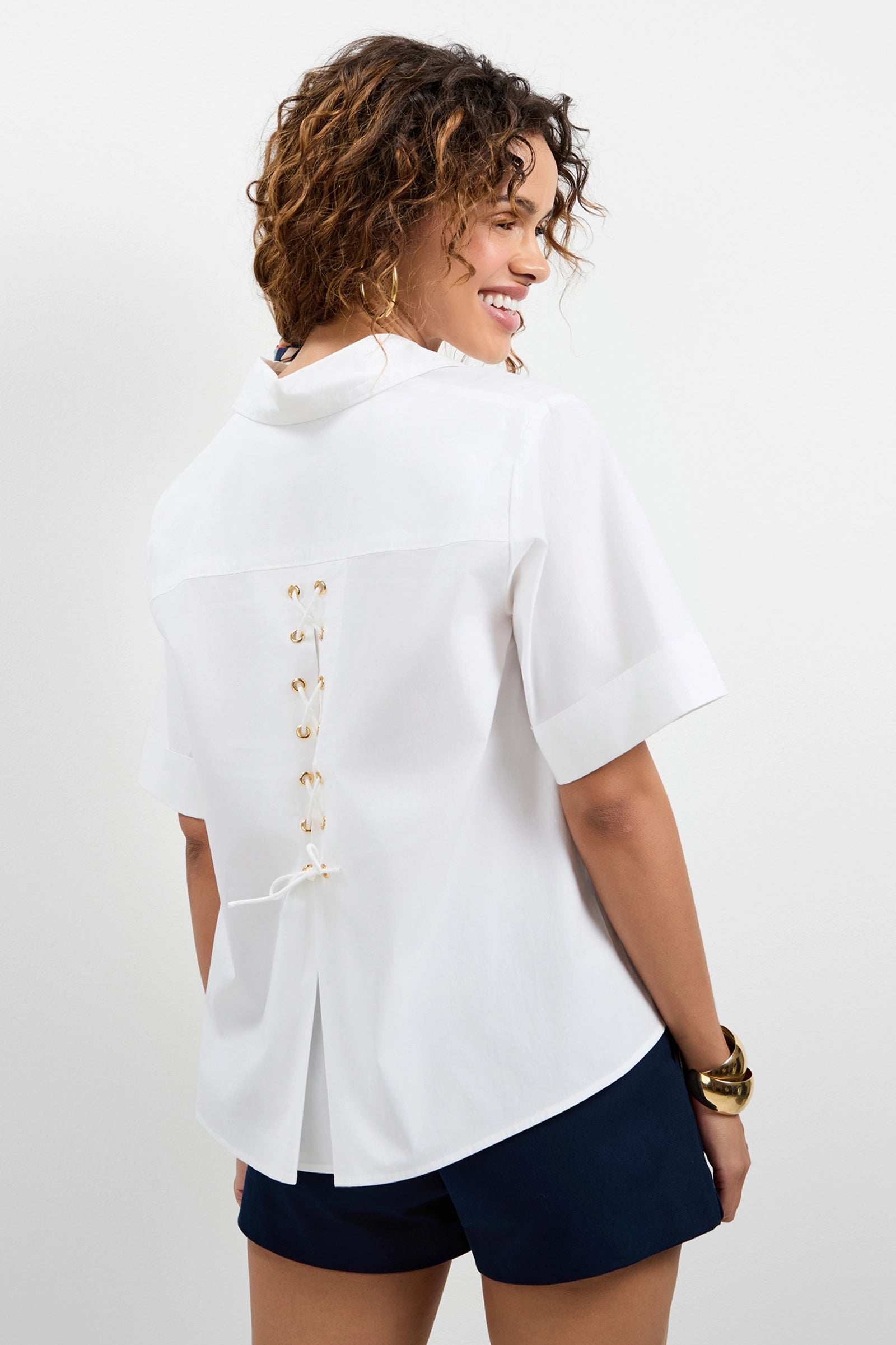 A woman smiles in the Triennale Lace-Up Back Top, a short-sleeve white blouse with gold eyelet lace-up detailing on the back, paired with dark navy shorts against a plain white background.