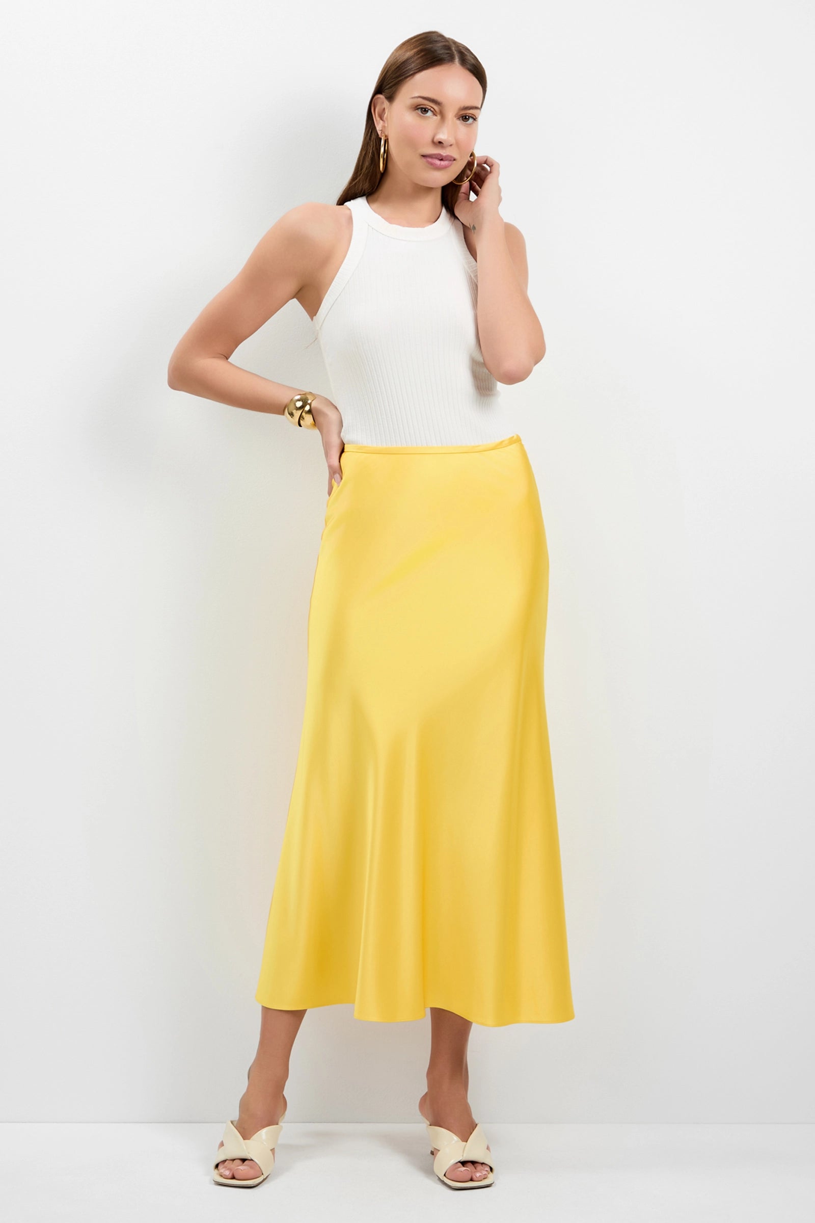 A woman with long, straight hair and a neutral expression poses against a white background in a sleeveless white top, the Rabina Flared Skirt in Lambretta Lemon, white open-toe heels, gold hoop earrings, and a gold bracelet.