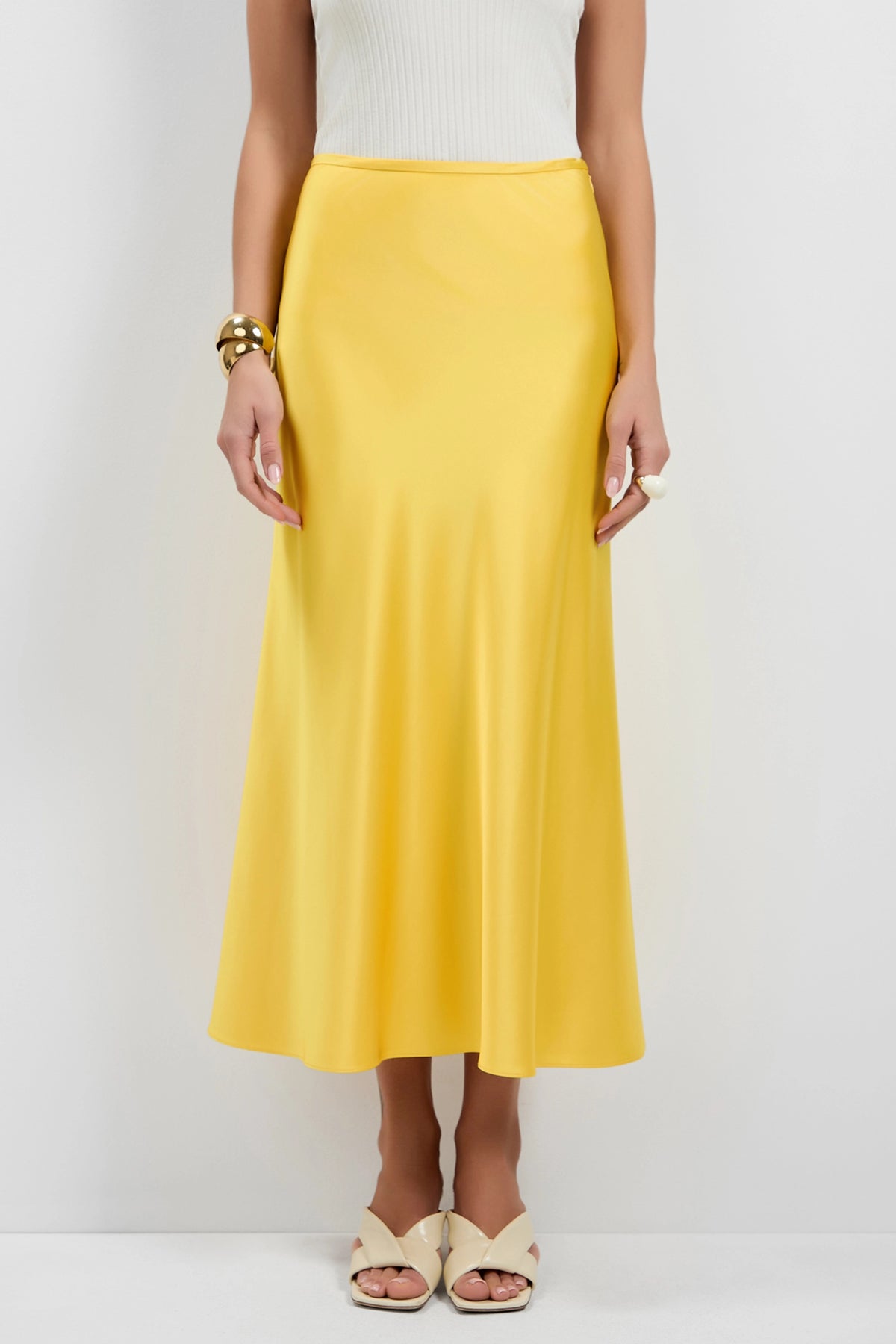 A woman, shown from the shoulders down, stands against a white background wearing a white sleeveless top, beige sandals, gold bracelets, and the Rabina Flared Skirt in yellow satin.
