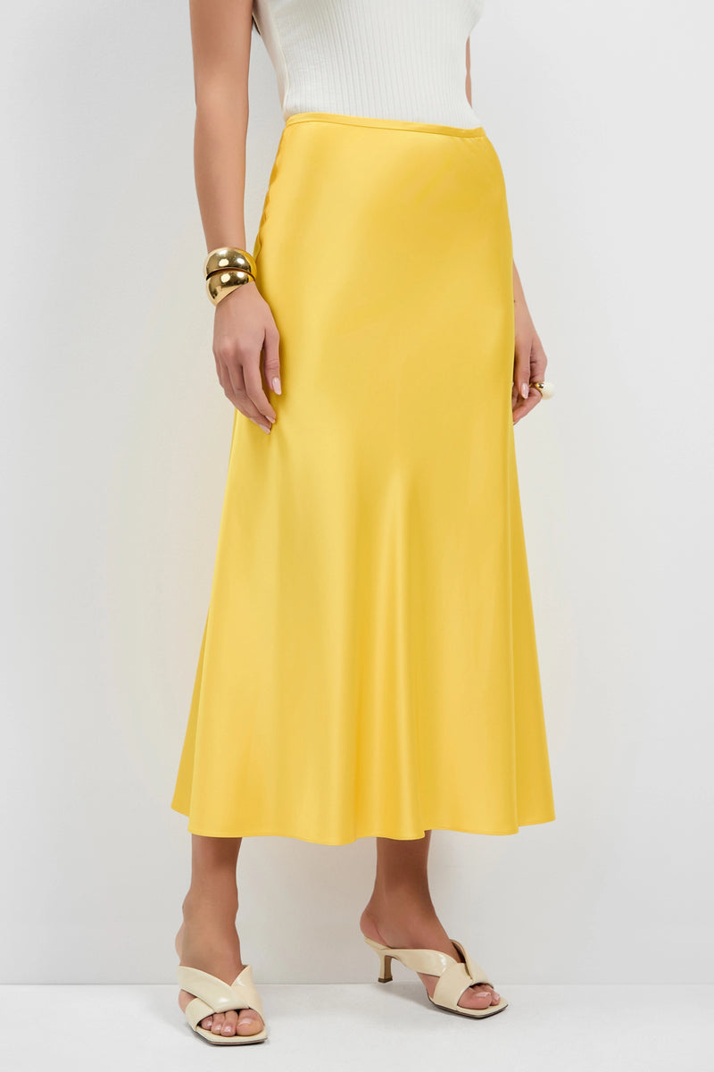 A woman, face not visible, stands against a white background wearing the Rabina Flared Skirt in shiny yellow, paired with a white sleeveless top, cream heeled sandals, and gold bracelets.