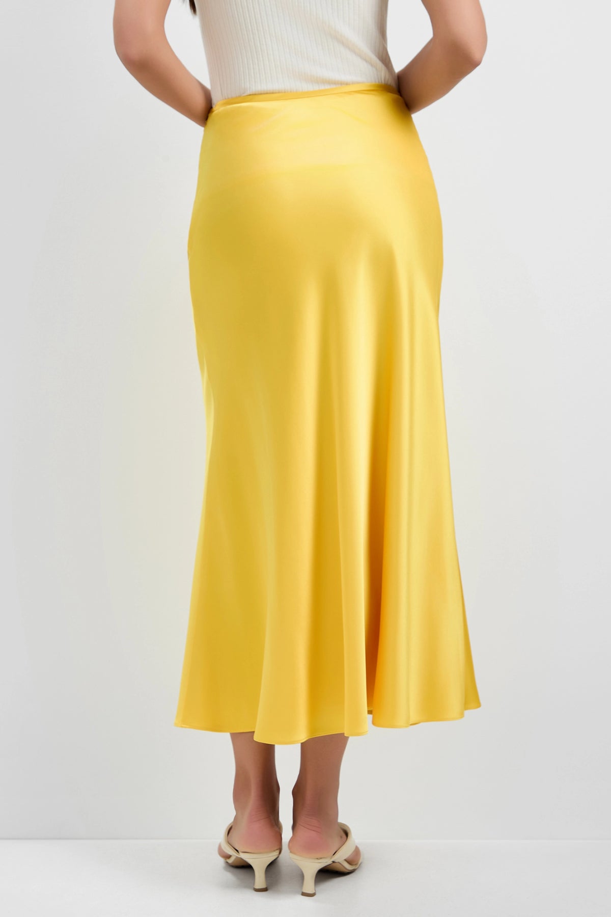 A woman wearing a white sleeveless top and the Rabina Flared Skirt in yellow satin stands with her back to the camera, paired with light heeled sandals against a plain white background.