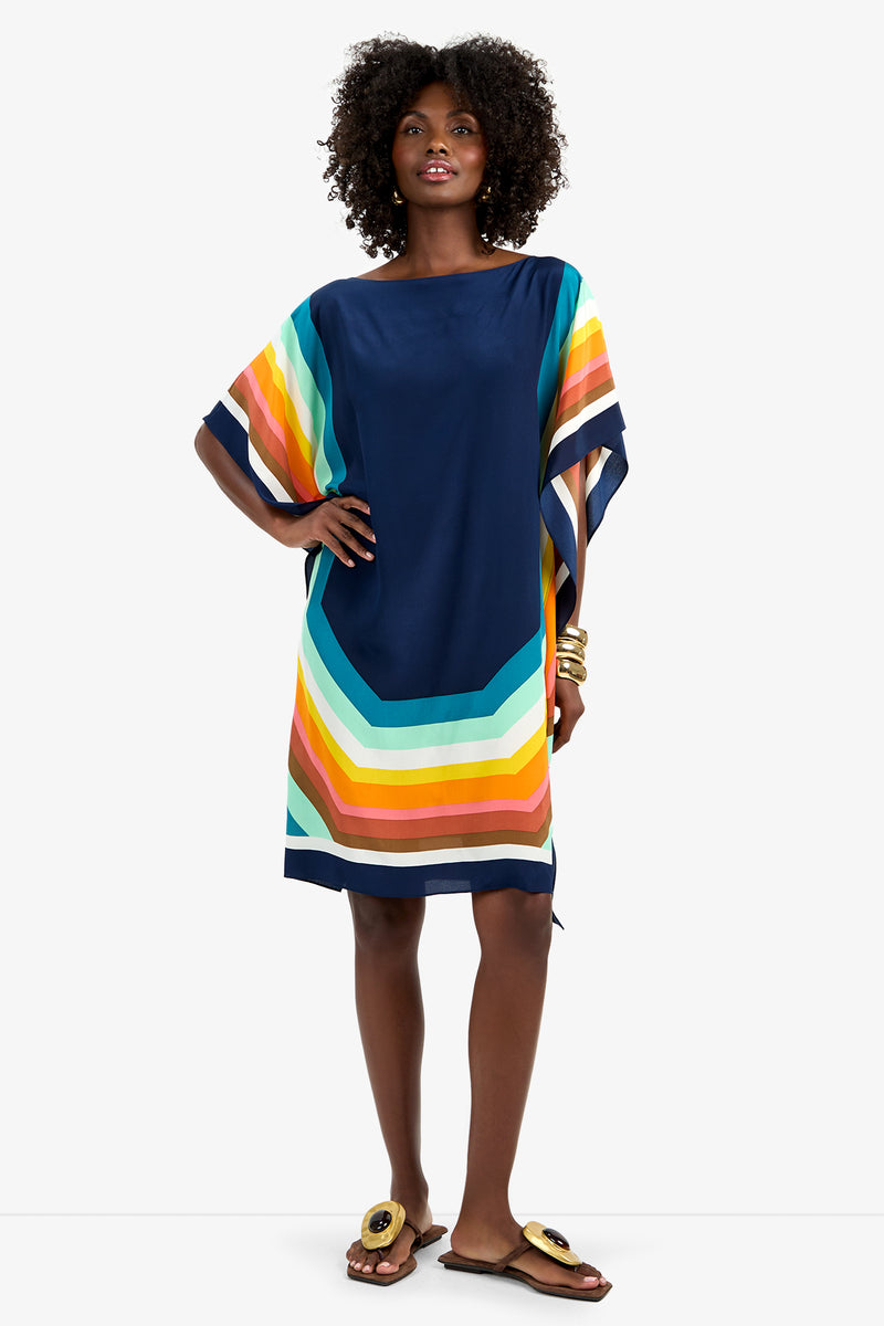 A woman wears Theodora Caftan Dress - Siena, a loose, knee-length navy dress featuring bold orange, yellow, teal, and white stripes on the sleeves and hem. She pairs it with curly hair, gold bracelets, and buckle-detailed sandals.