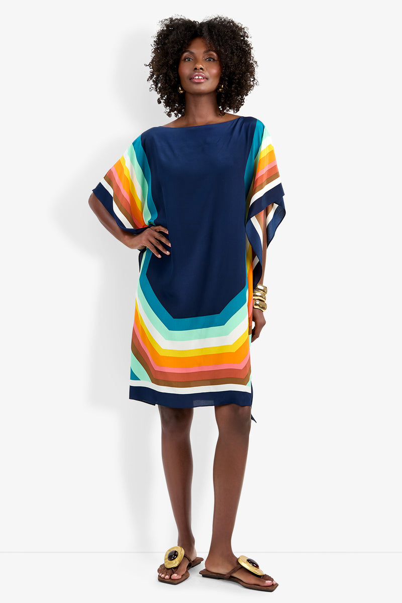 A woman stands against a white background wearing the Theodora Caftan Dress, a loose-fitting, knee-length piece with wide, colorful diagonal stripes. She completes the look with sandals and gold bracelets.