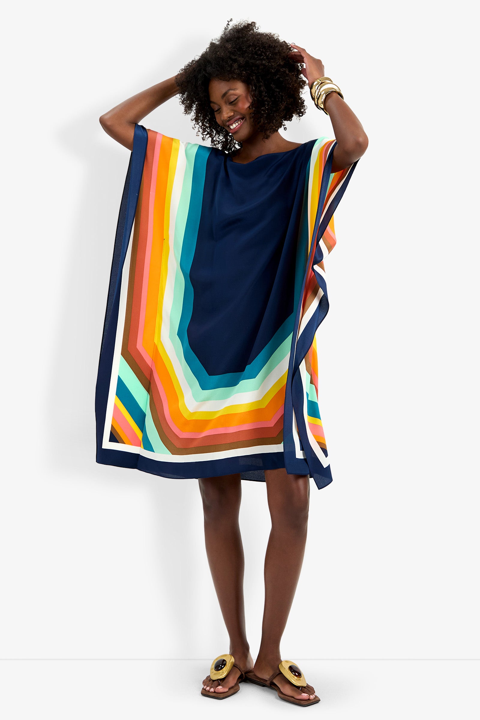 A woman smiles in Theodora Caftan Dress, featuring flowy fabric and bold geometric stripes in navy, orange, yellow, green, and white. She completes the look with sandals and bangles, posing with a hand in her hair.