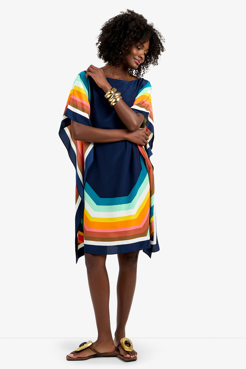 A woman stands smiling against a white background, wearing the Theodora Caftan Dress - Siena, a loose navy blue dress with bold, colorful striped edges, paired with gold bracelets and sandals.
