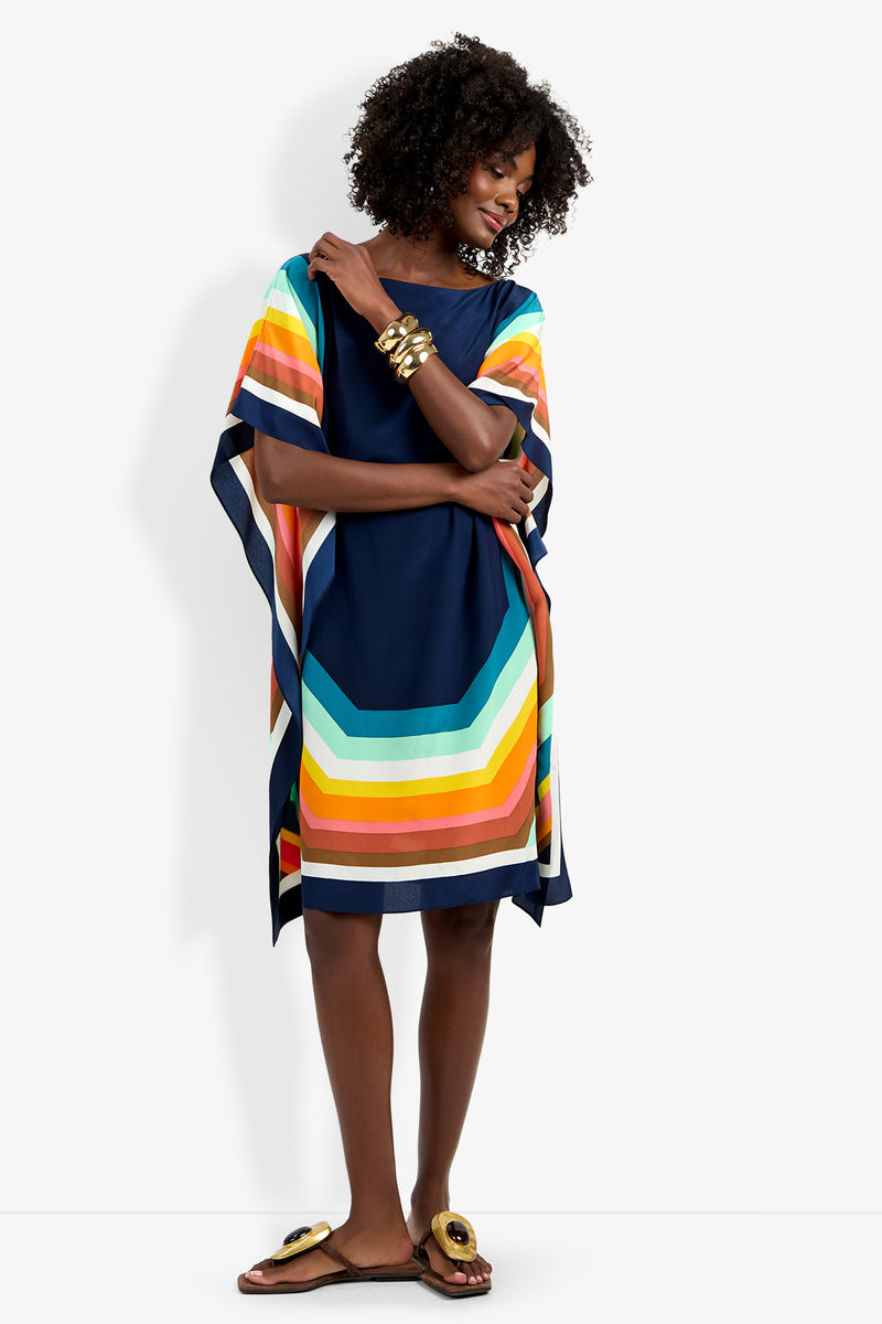 A woman in the Theodora Caftan Dress, a loose navy blue dress with bold colorful stripes, smiles against a plain white background. She accessorizes with gold bangles and sandals, and has curly hair.