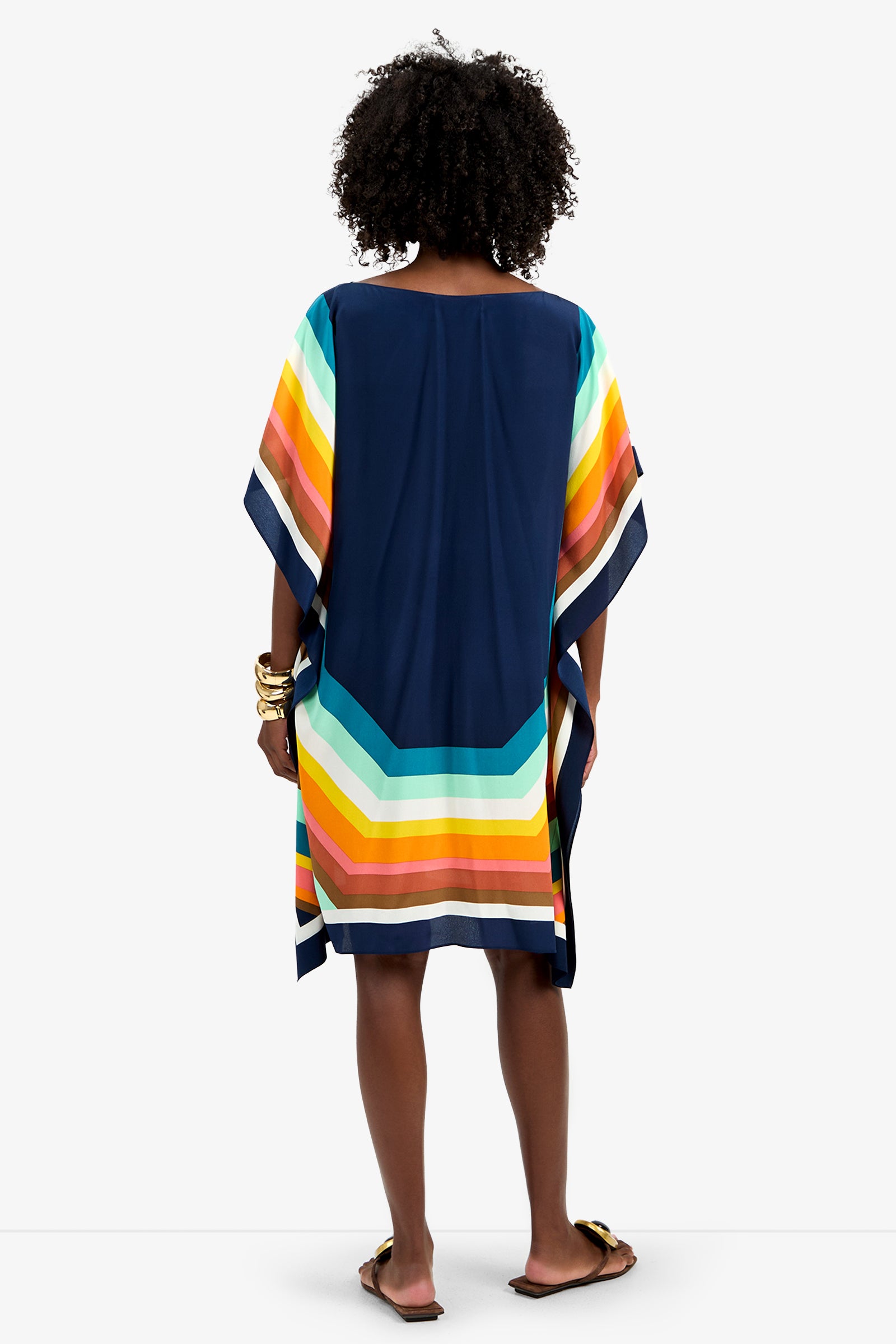 A woman with curly hair faces away, wearing Theodora Caftan Dress - Siena, a loose navy dress featuring wide, colorful horizontal stripes in yellow, orange, green, and blue at the hem and sleeves. She pairs it with sandals and gold bangles.