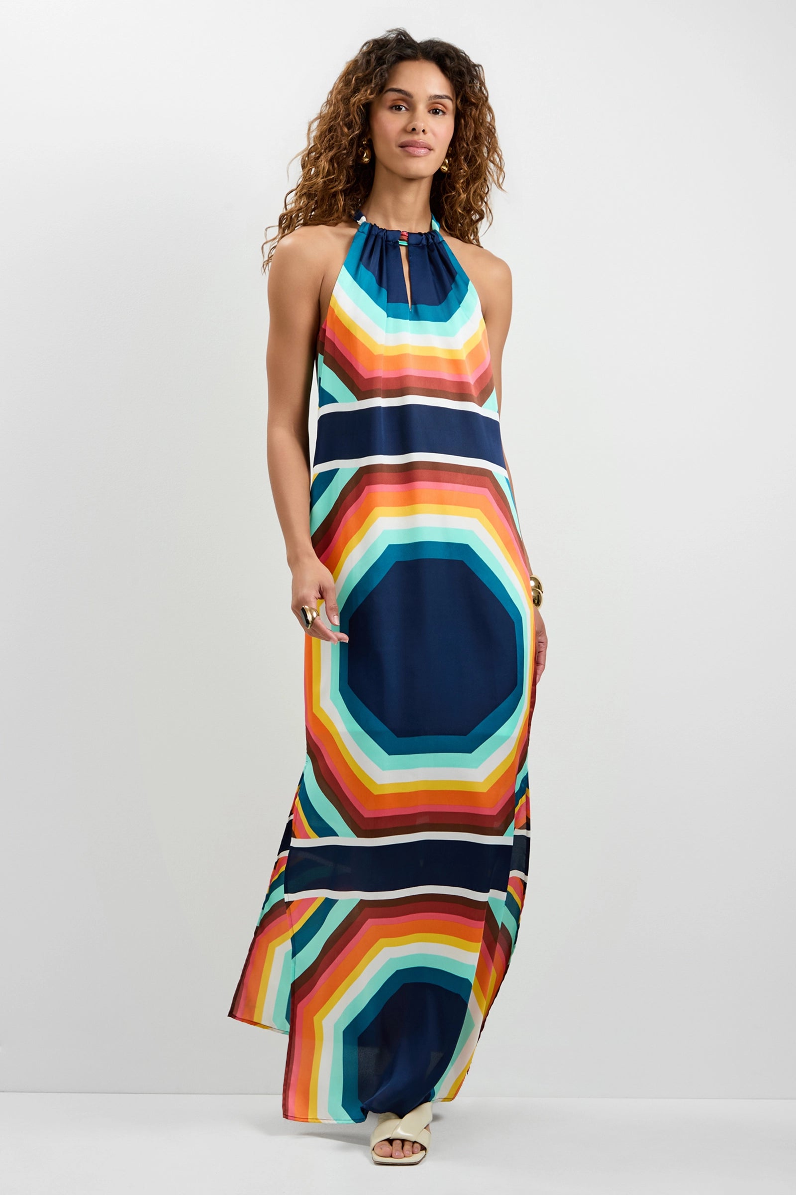 A woman with curly hair smiles slightly while wearing the Emilo Sleeveless Maxi Dress, which features a bold, colorful geometric pattern. She stands against a plain white background.