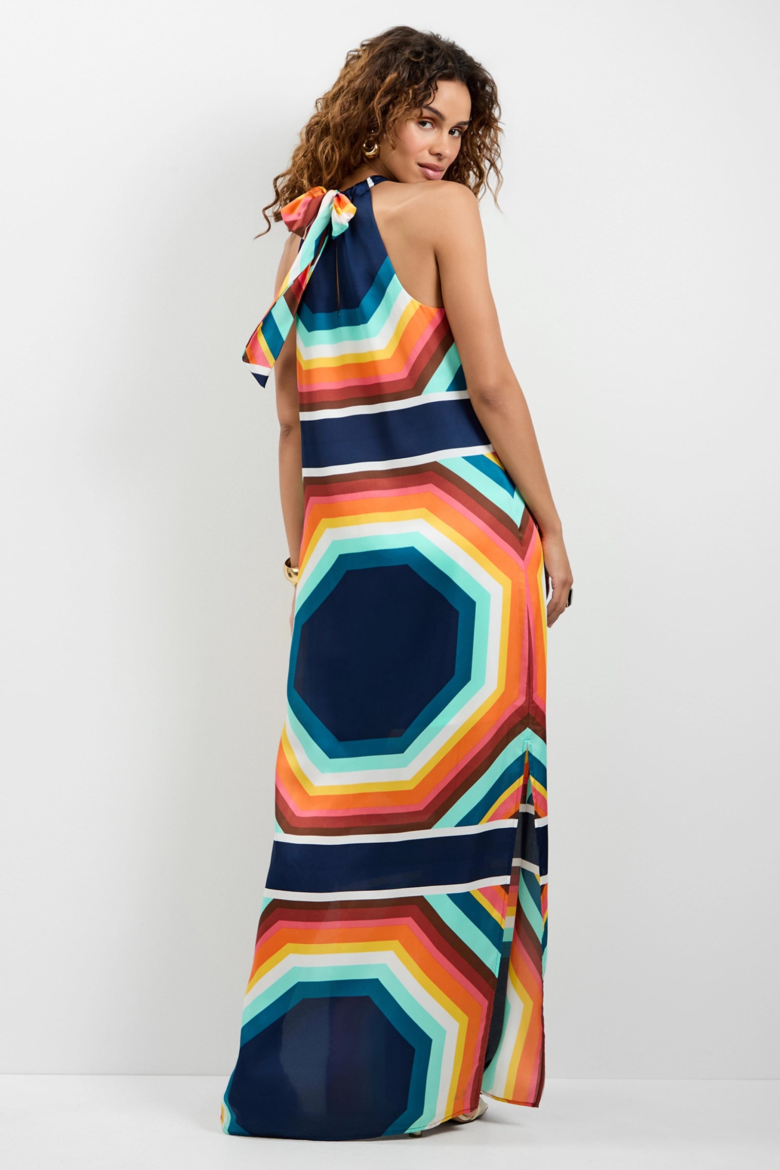 A woman faces away wearing the Emilo Sleeveless Maxi Dress, a floor-length style with bold geometric patterns in orange, blue, yellow, and teal. A bow decorates one shoulder as she glances back over her shoulder.