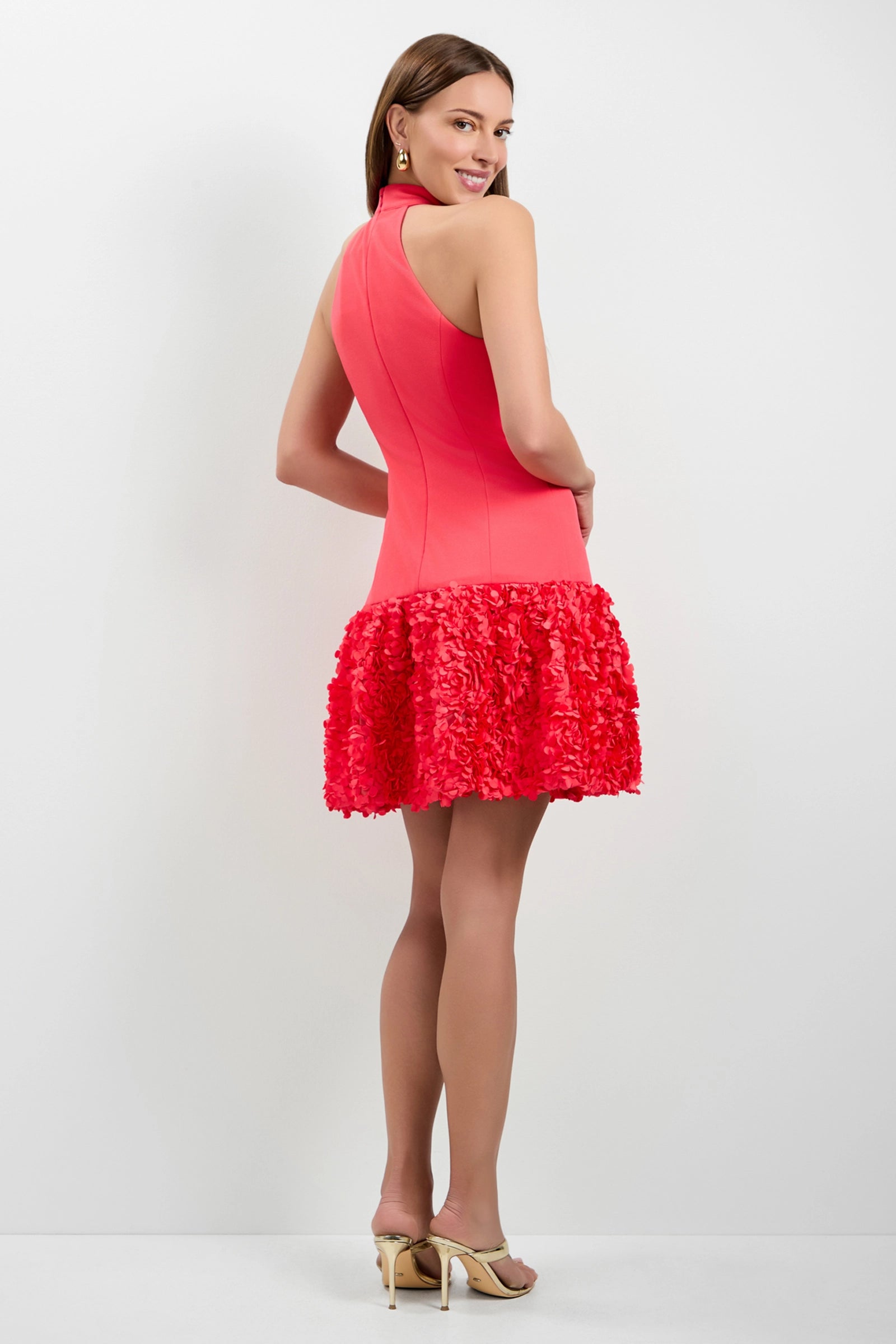 A woman models the Tibaldi 3D Floral Mini Dress, a sleeveless bright pink dress with a textured ruffled skirt. She pairs it with beige heels and gold earrings, smiling over her shoulder against a plain white background.