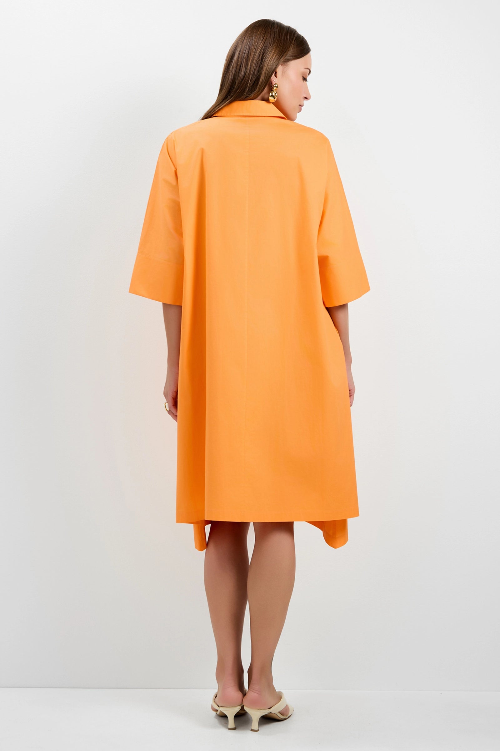 A woman with long brown hair stands facing away, wearing the Mirabello Shirt Dress—an orange, loose-fitting, knee-length style with elbow-length sleeves—and cream-colored heeled sandals against a plain white background.