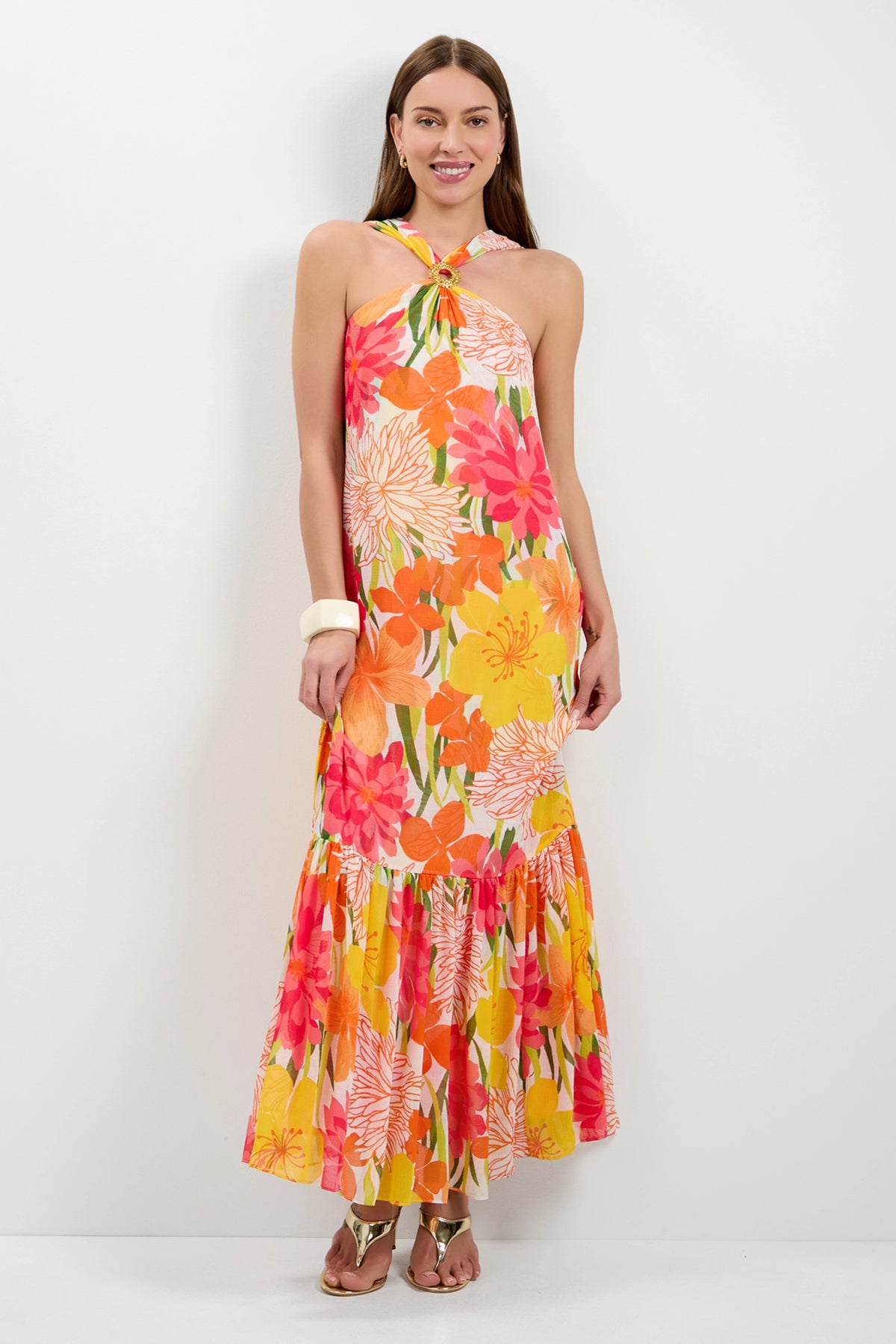 A woman smiles against a white wall, wearing the Navigli Sleeveless Maxi Dress in MULTI—a colorful halter-neck maxi with a bold floral pattern. She has long hair, sandals, and accessorizes with a white bangle.