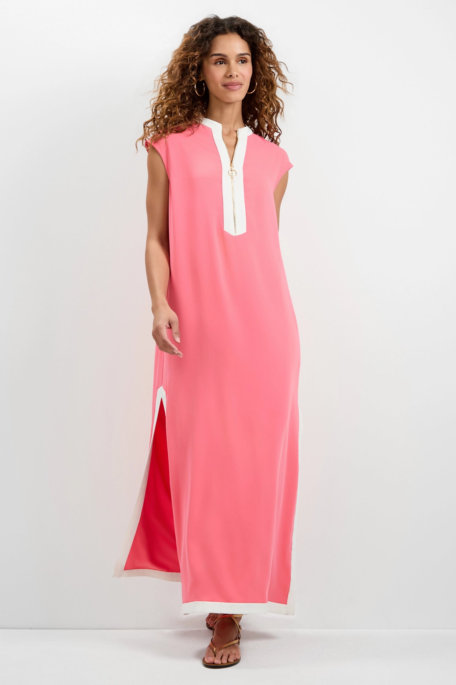 A woman with curly hair smiles while posing in the Nuvola Zip-Front Midi Dress—a sleeveless, ankle-length pink dress with white trim and a front slit—against a plain white background, one hand slightly raised.