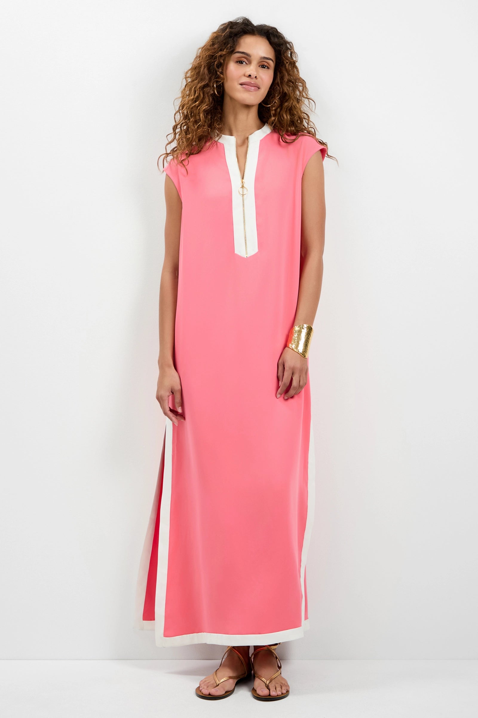 A woman with curly hair wearing the Nuvola Zip-Front Midi Dress—sleeveless, ankle-length pink with white trim—plus a gold bracelet and sandals, stands against a white background, looking forward and smiling slightly.