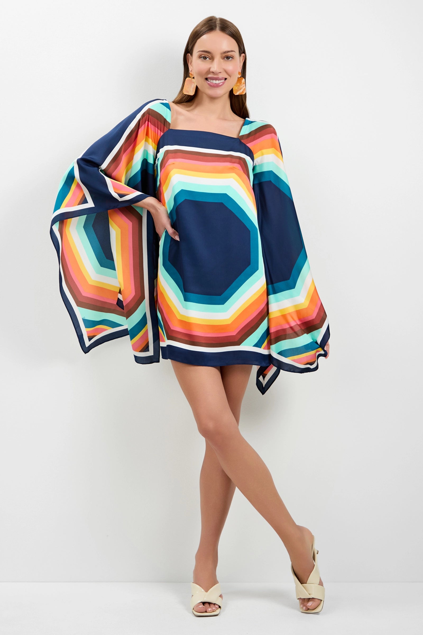 A woman smiles and poses in the Aqualane Side Flounce Dress, featuring colorful geometric patterns, wide flowing sleeves, and neutral heels, against a plain white background.