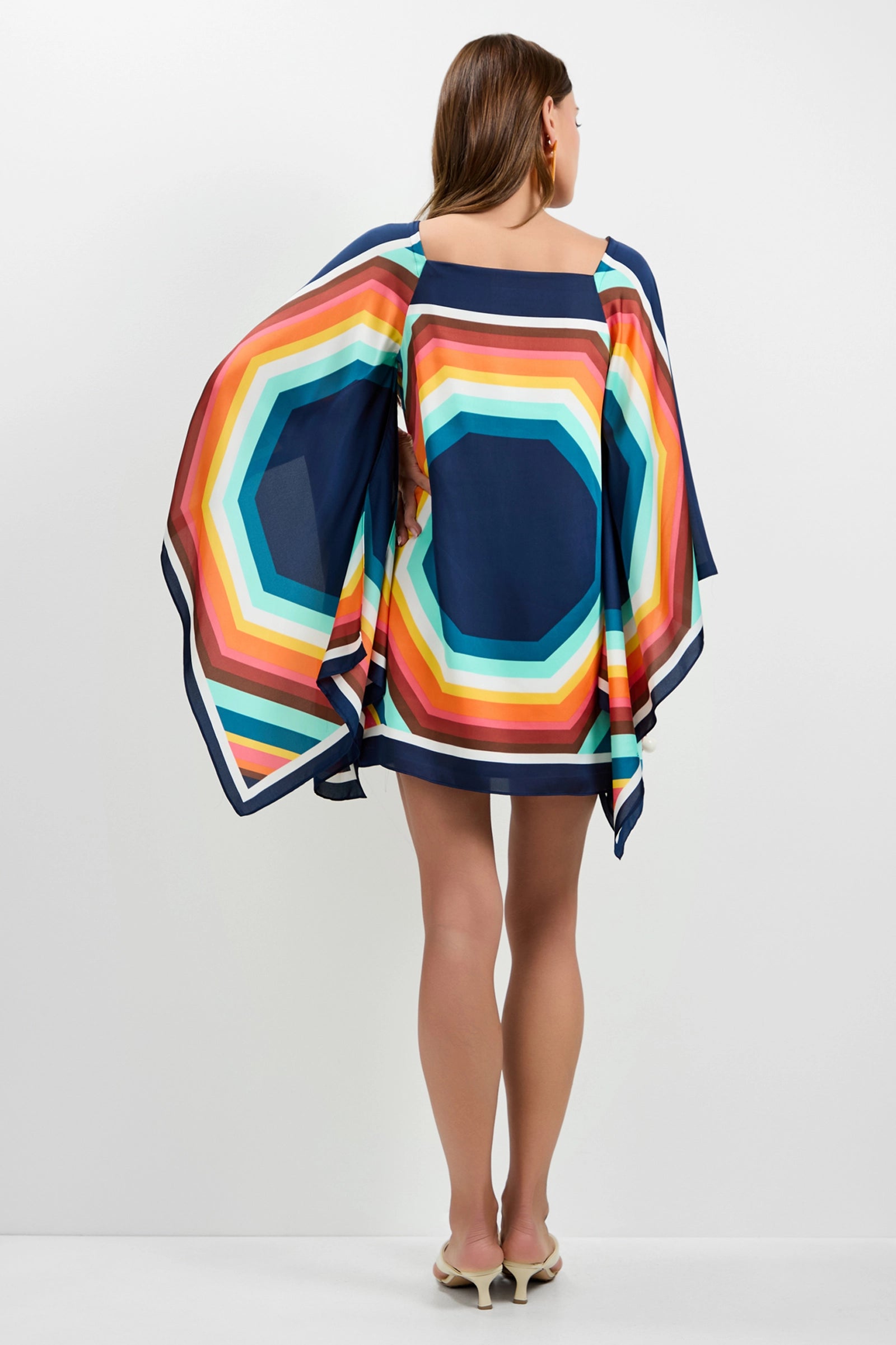 A woman with shoulder-length brown hair faces away, wearing the Aqualane Side Flounce Dress—a vibrant, geometric-patterned dress in blue, orange, yellow, and green tones with wide sleeves—paired with light-colored heels.
