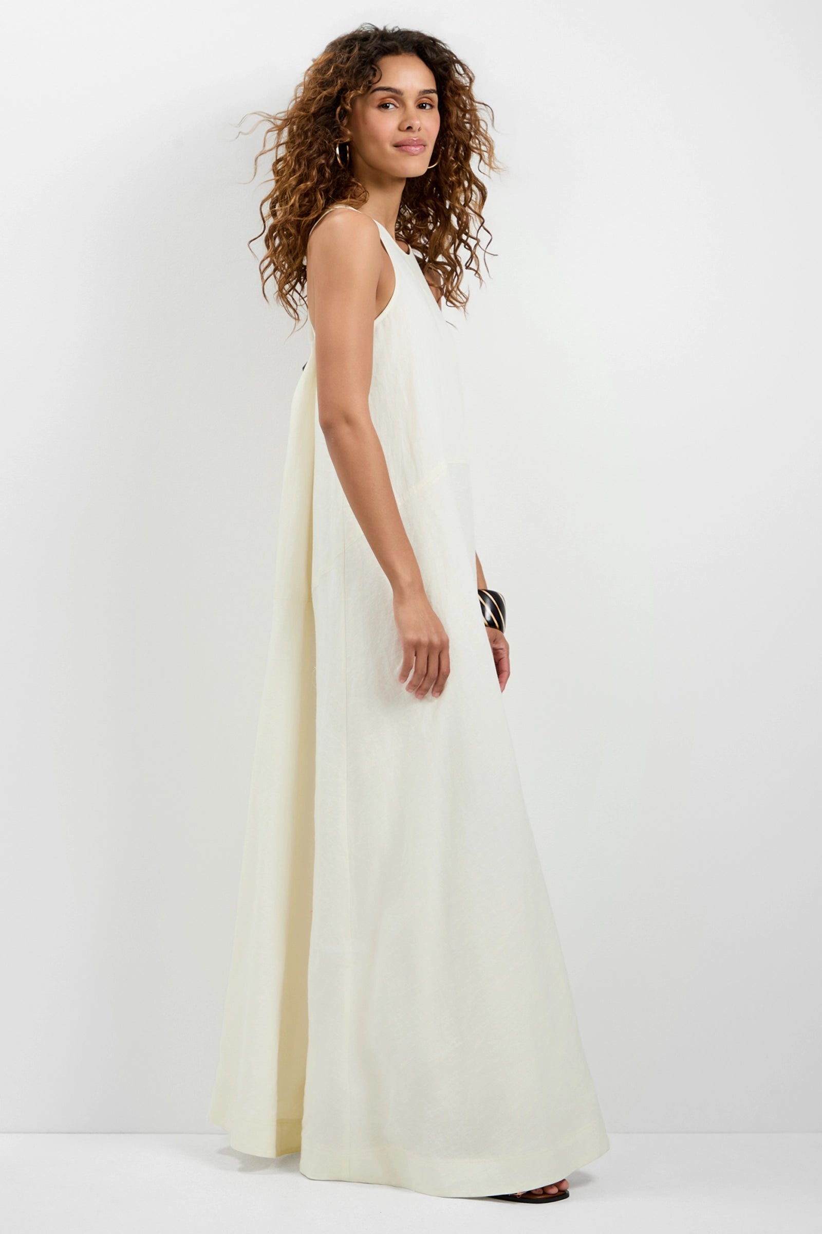 A woman with curly hair stands sideways against a white background wearing the Pichi 2 Maxi Dress, a long, sleeveless, loose-fitting cream dress paired with sandals. She glances toward the camera with a relaxed expression.