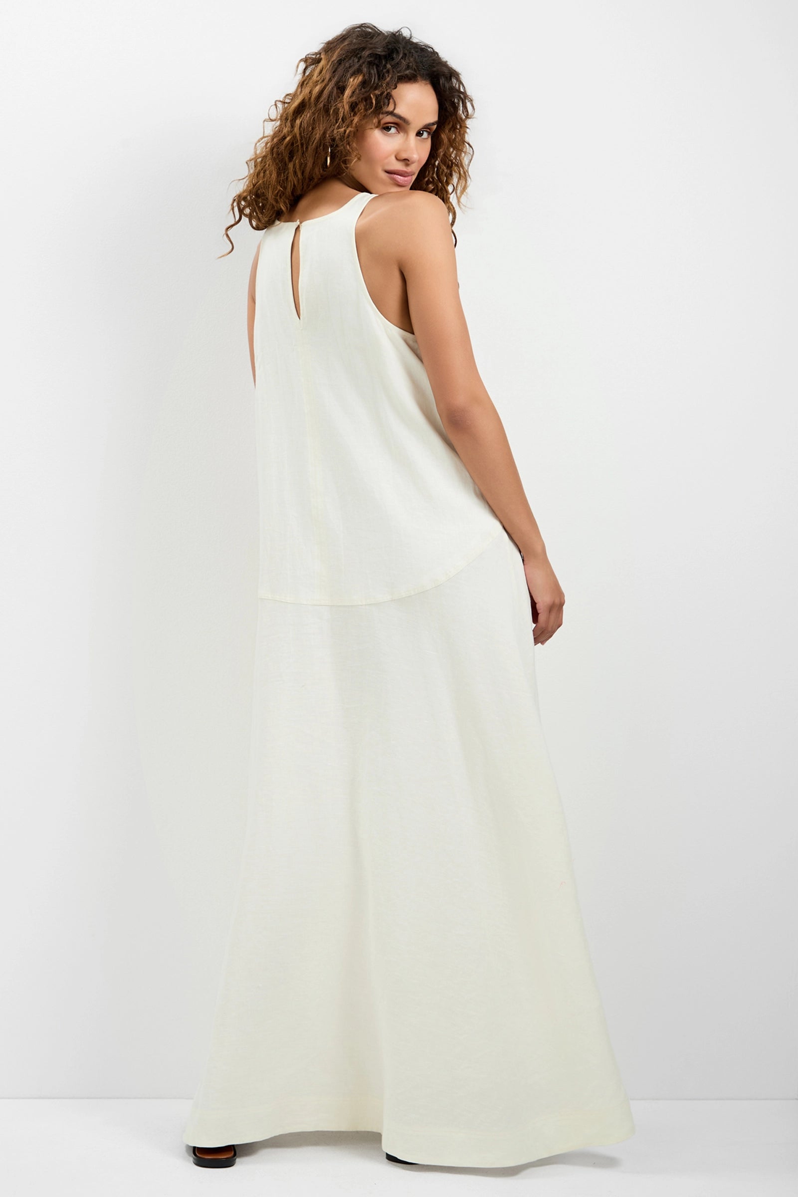A woman with curly hair stands with her back to the camera, looking over her shoulder while wearing the Pichi 2 Maxi Dress—a sleeveless, long, white dress—against a plain white background.