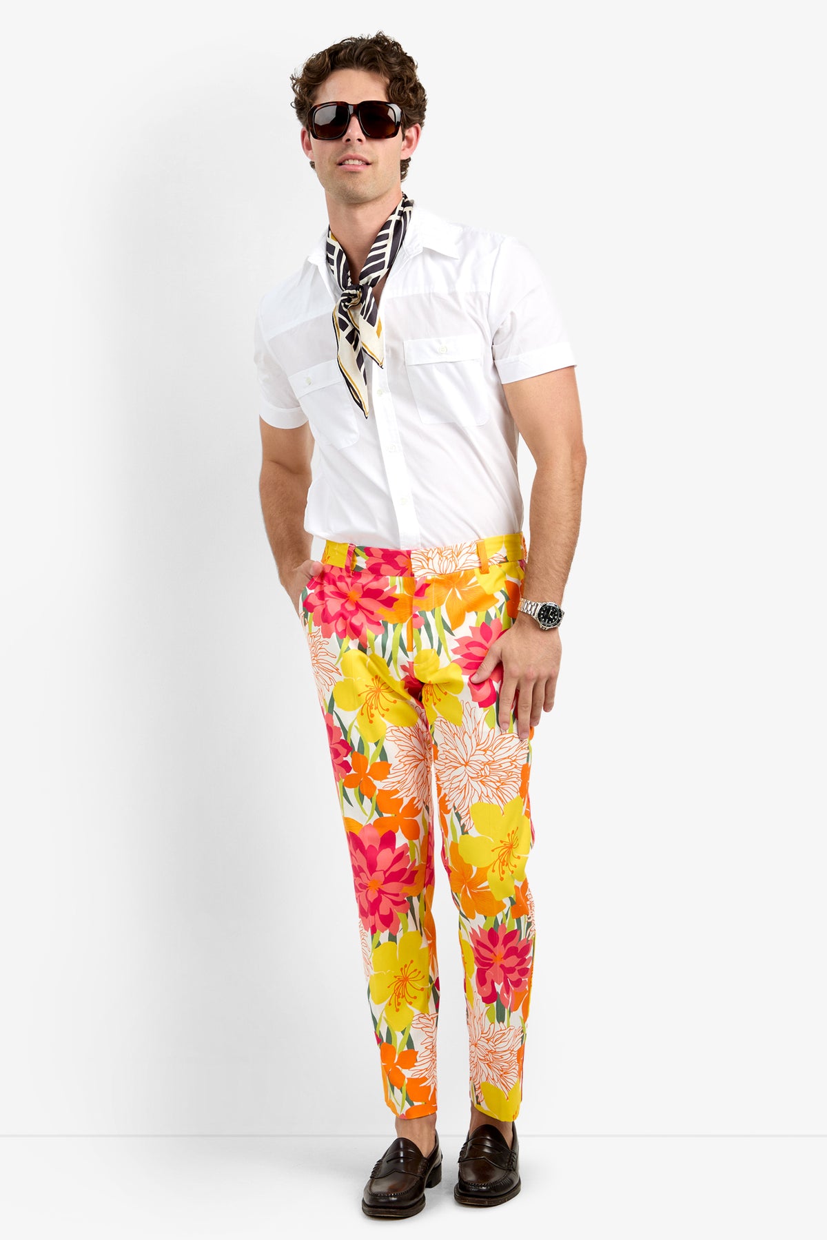 A man stands against a white background wearing the Clyde Slim Trouser - Como Herringbone, paired with a white short-sleeve shirt, black-and-white scarf, sunglasses, brown shoes, a watch, and bright floral pants in yellow, pink, and orange.
