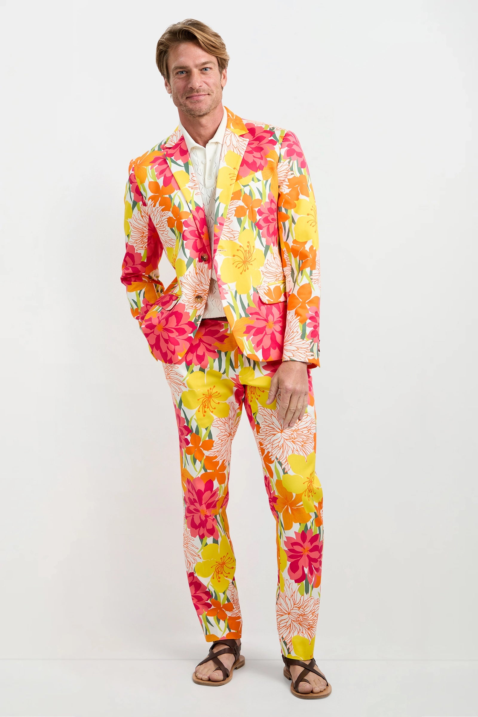 A man stands against a white background wearing the Clyde Slim Trouser - Como Herringbone in MULTI, styled with a white shirt and brown sandals. He smiles slightly, one hand in his pocket, showcasing the vibrant, patterned trousers.