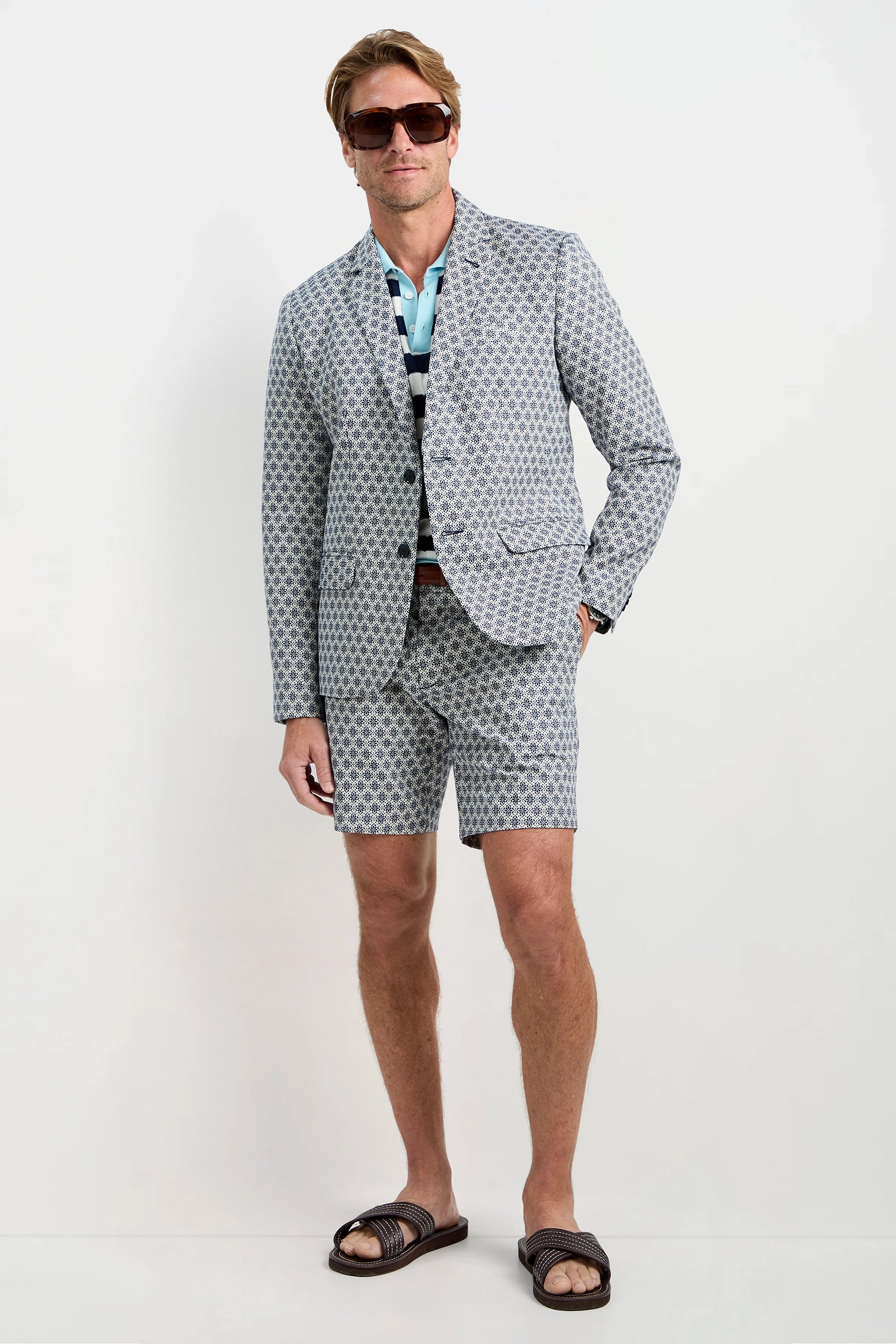 A man stands against a white background wearing the Thurston Blazer - Daisy Jacquard with matching shorts, a striped shirt, sunglasses, and dark sandals.
