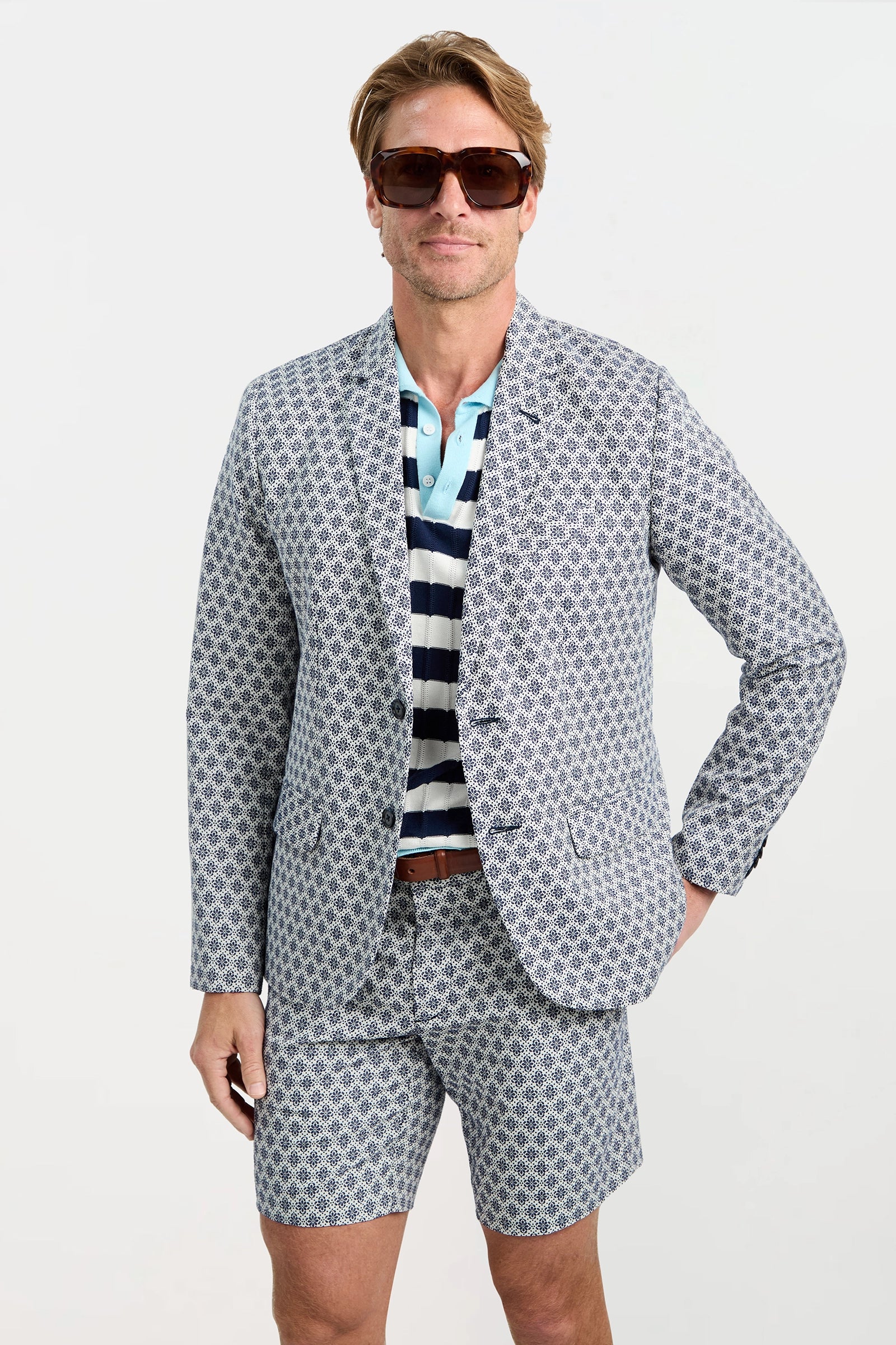 A man wearing the Thurston Blazer - Daisy Jacquard with matching shorts, a striped polo, sunglasses, and a brown belt stands against a plain white background.