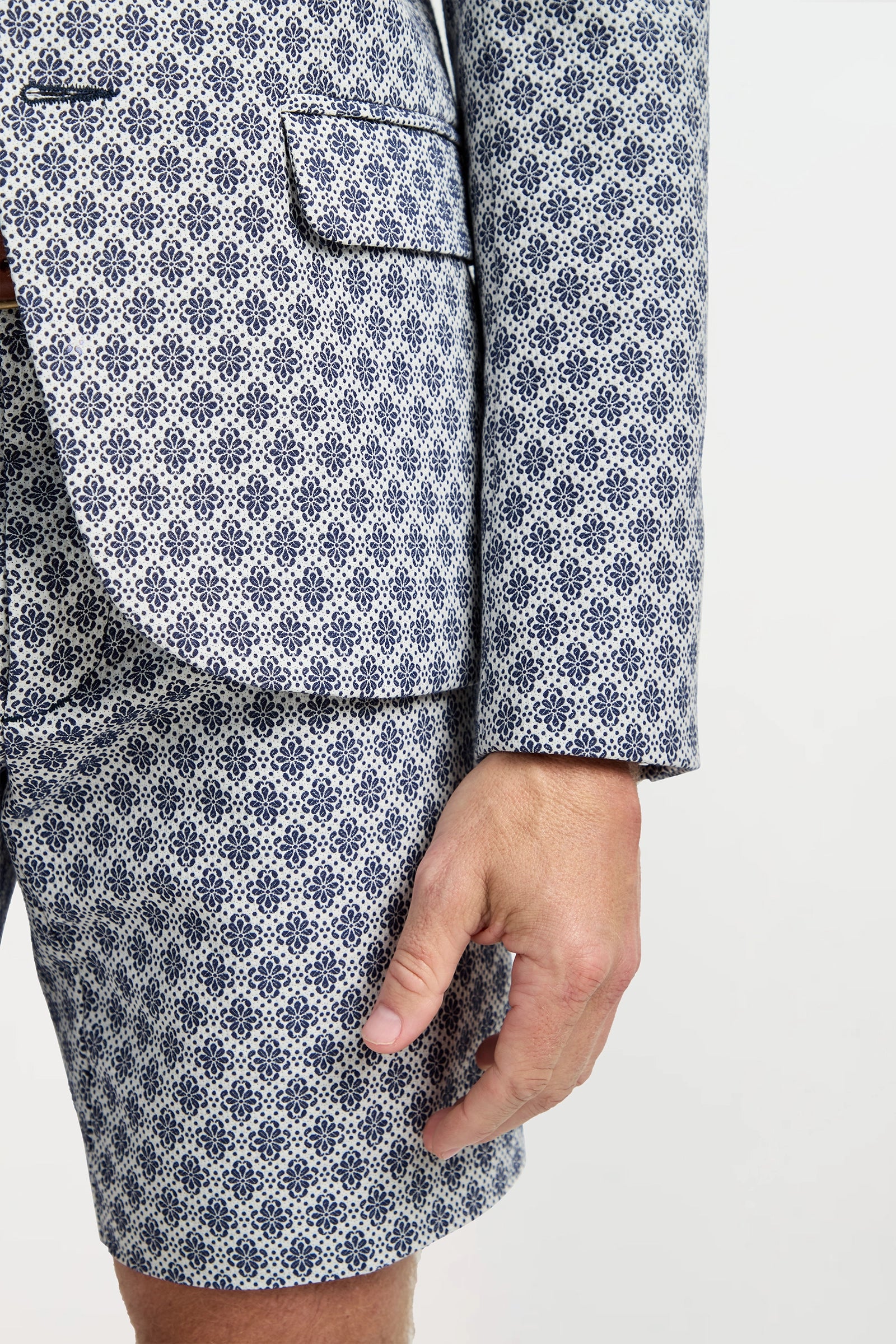 A person wearing the Thurston Blazer in Daisy Jacquard—a white and blue geometric patterned suit jacket and shorts—stands against a plain background with their right arm by their side.