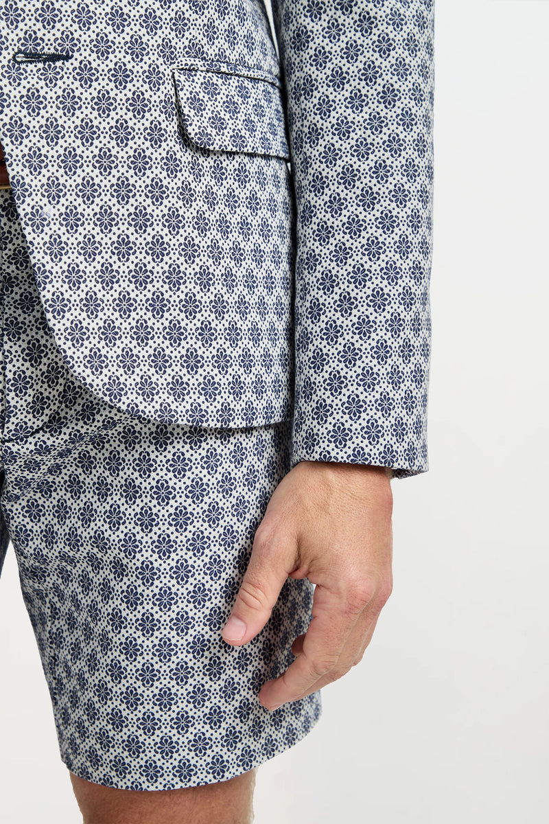 A person wearing the Thurston Blazer in Daisy Jacquard—a white and blue geometric patterned suit jacket and shorts—stands against a plain background with their right arm by their side.