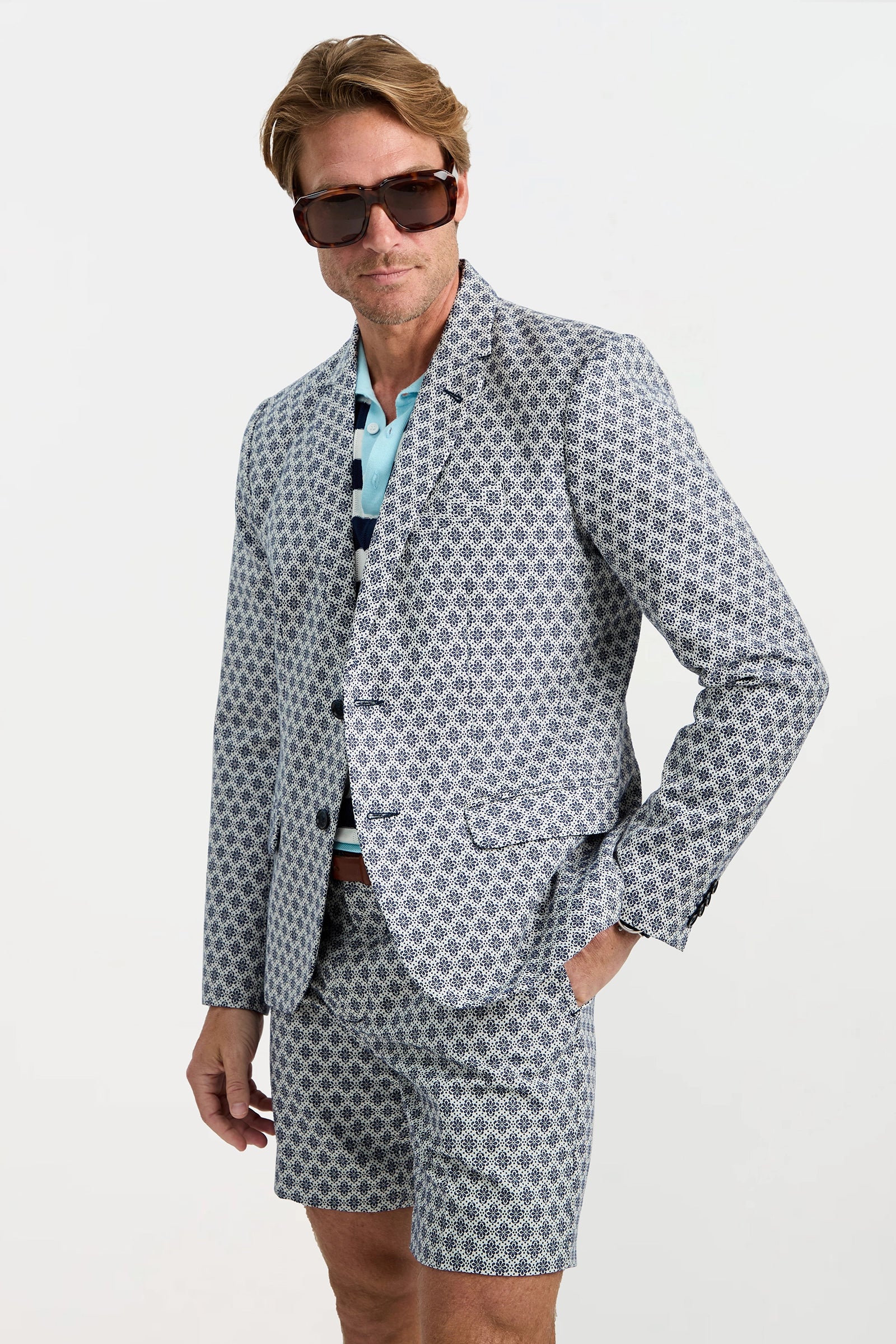 A man wears the Thurston Blazer in Daisy Jacquard, matching shorts, a blue striped polo shirt, and large dark sunglasses, standing with one hand in his pocket against a white background.