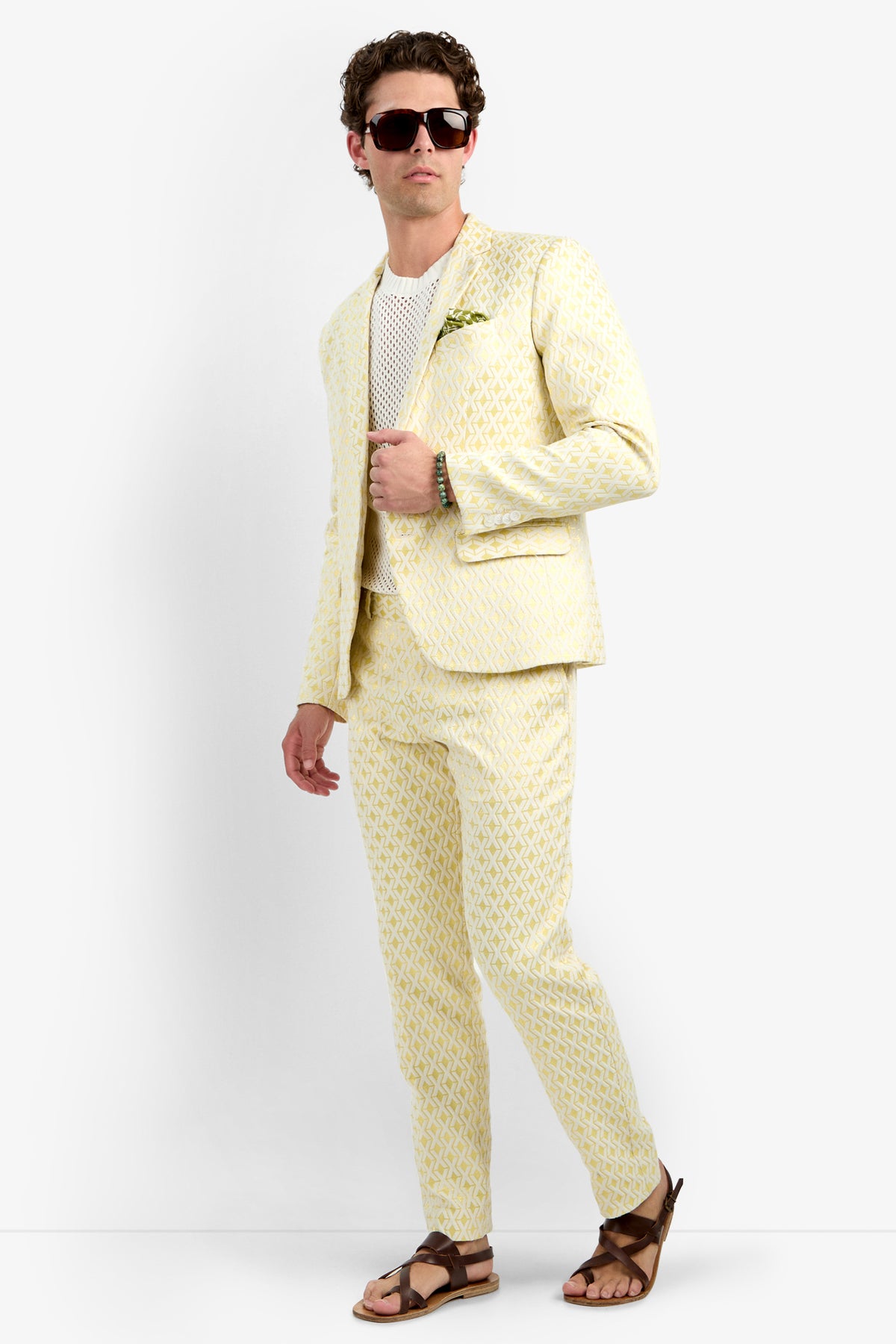 A man stands against a white background in the Thurston Blazer - Crossroads Jacquard, light yellow patterned suit, white mesh shirt, brown sandals, dark sunglasses, with a green pocket square and one hand in his pocket.