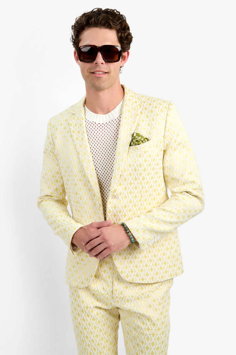 A man in the Thurston Blazer - Crossroads Jacquard, styled with a green pocket square, white mesh shirt, dark sunglasses, and a bracelet, stands against a plain white background, smiling slightly.