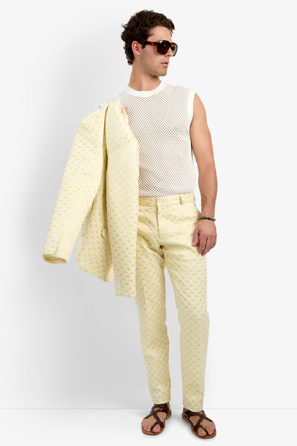 A man holds the Thurston Blazer - Crossroads Jacquard over his shoulder, wearing brown sunglasses, a sleeveless white mesh shirt, yellow patterned pants, and brown sandals while looking to the side.