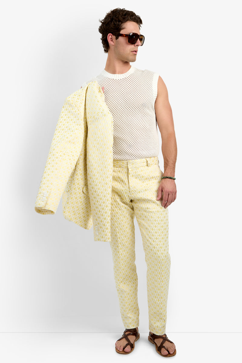 A man holds the Thurston Blazer - Crossroads Jacquard over his shoulder, wearing brown sunglasses, a sleeveless white mesh shirt, yellow patterned pants, and brown sandals while looking to the side.
