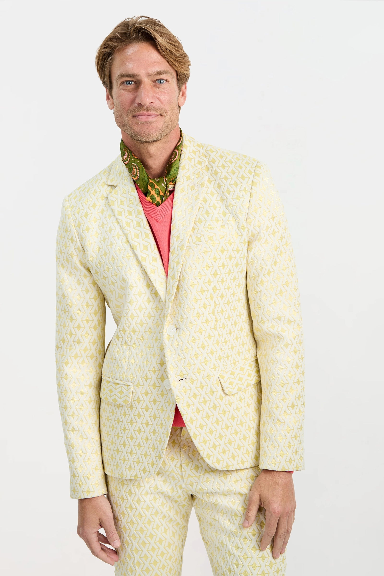 A man with light brown hair, smiling slightly, stands against a plain white background wearing the Thurston Blazer - Crossroads Jacquard, a coral pink shirt, and a green patterned scarf.