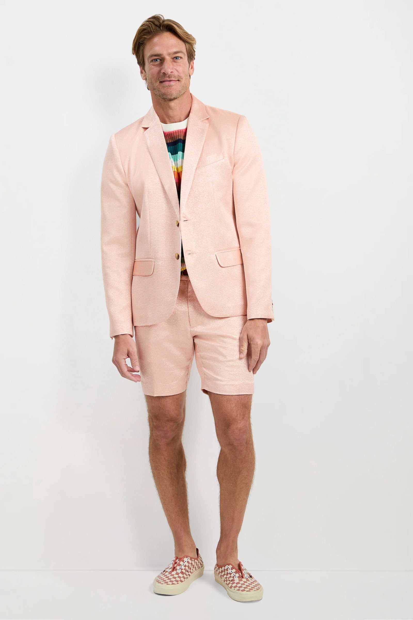 A man stands against a white background wearing the Thurston Blazer - Metallic Jacquard with matching shorts, a multicolored striped sweater, and checkered slip-on shoes. He smiles and keeps one hand by his side.