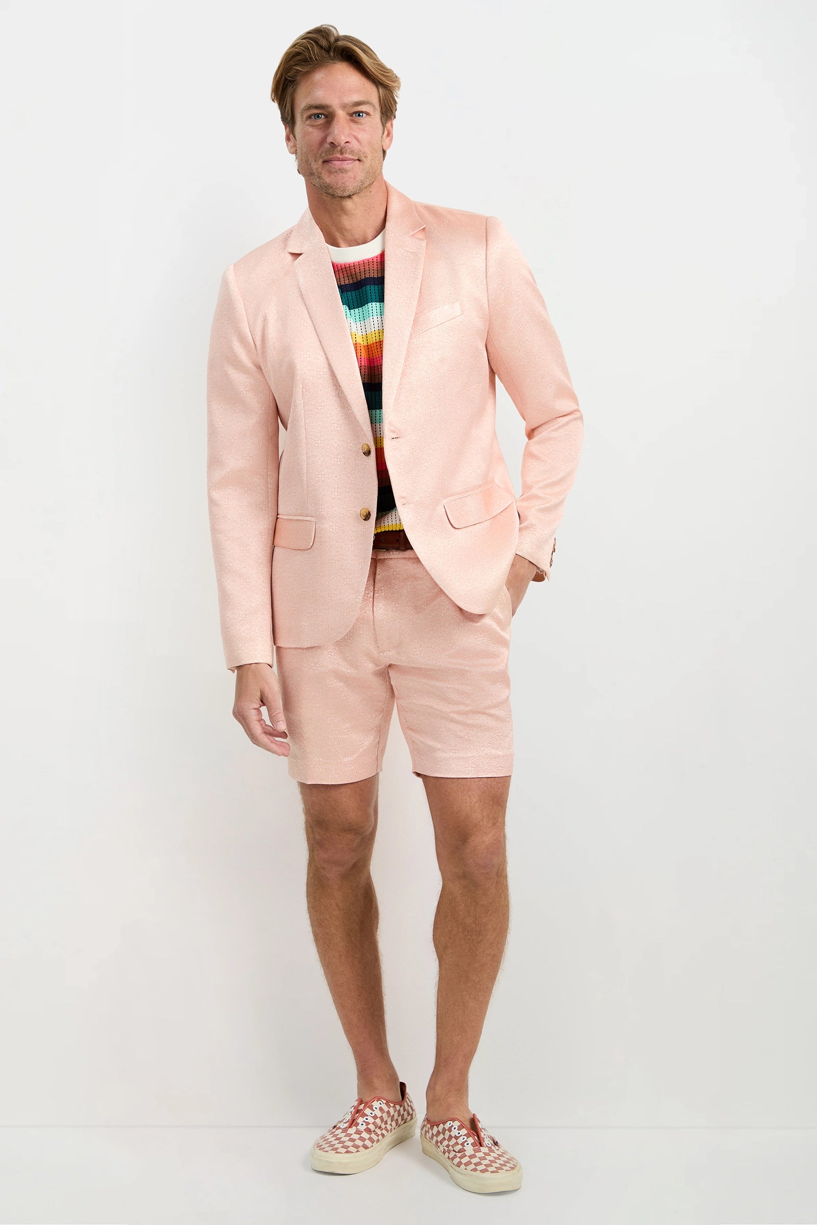 A man stands against a plain background wearing the Thurston Blazer in PALAZZO PINK metallic jacquard, paired with matching shorts, a colorful striped sweater, and red-and-white checkered slip-on shoes.