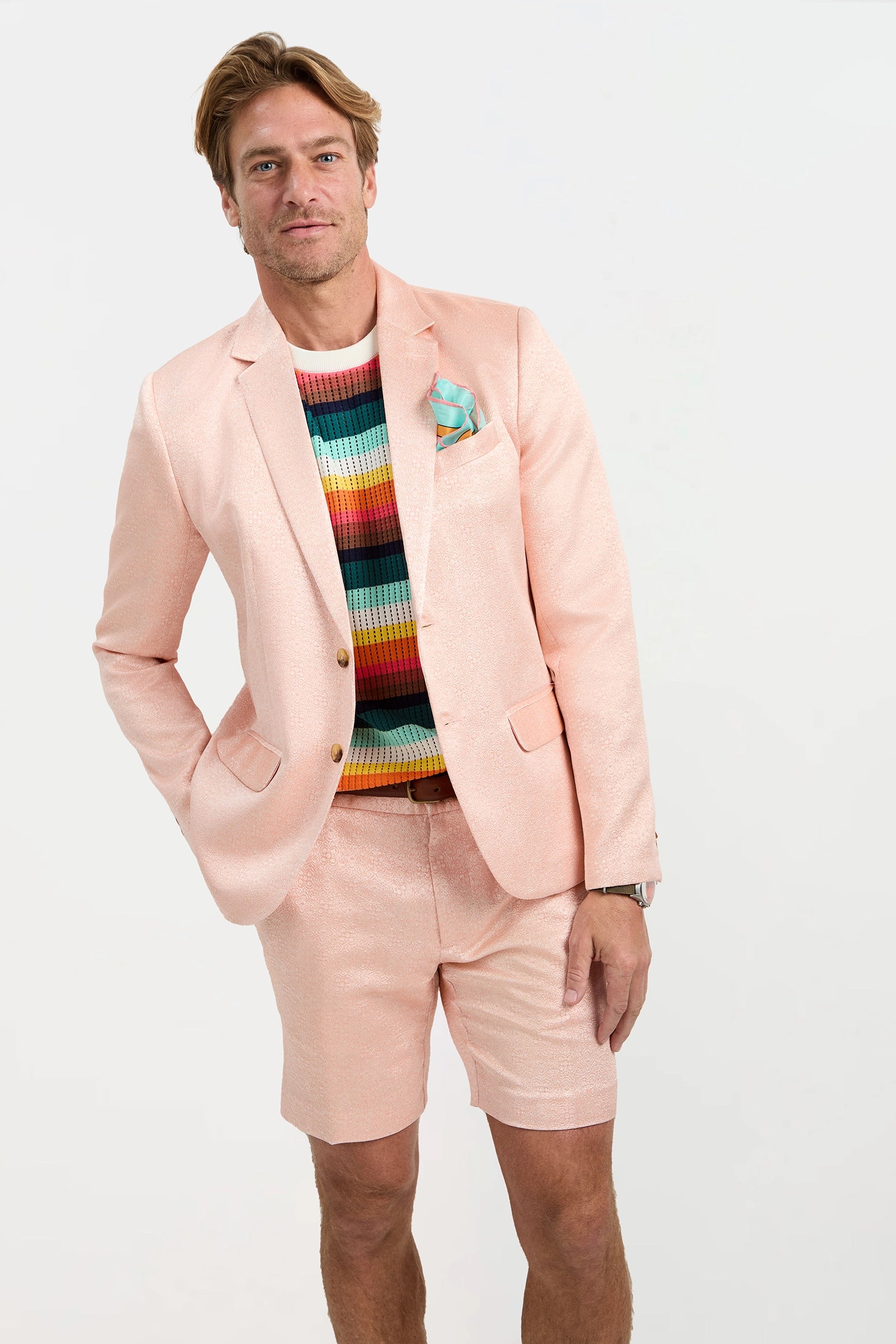 A man stands against a light background wearing the Thurston Blazer - Metallic Jacquard with matching shorts and a colorful striped sweater, one hand in his pocket and smiling slightly.
