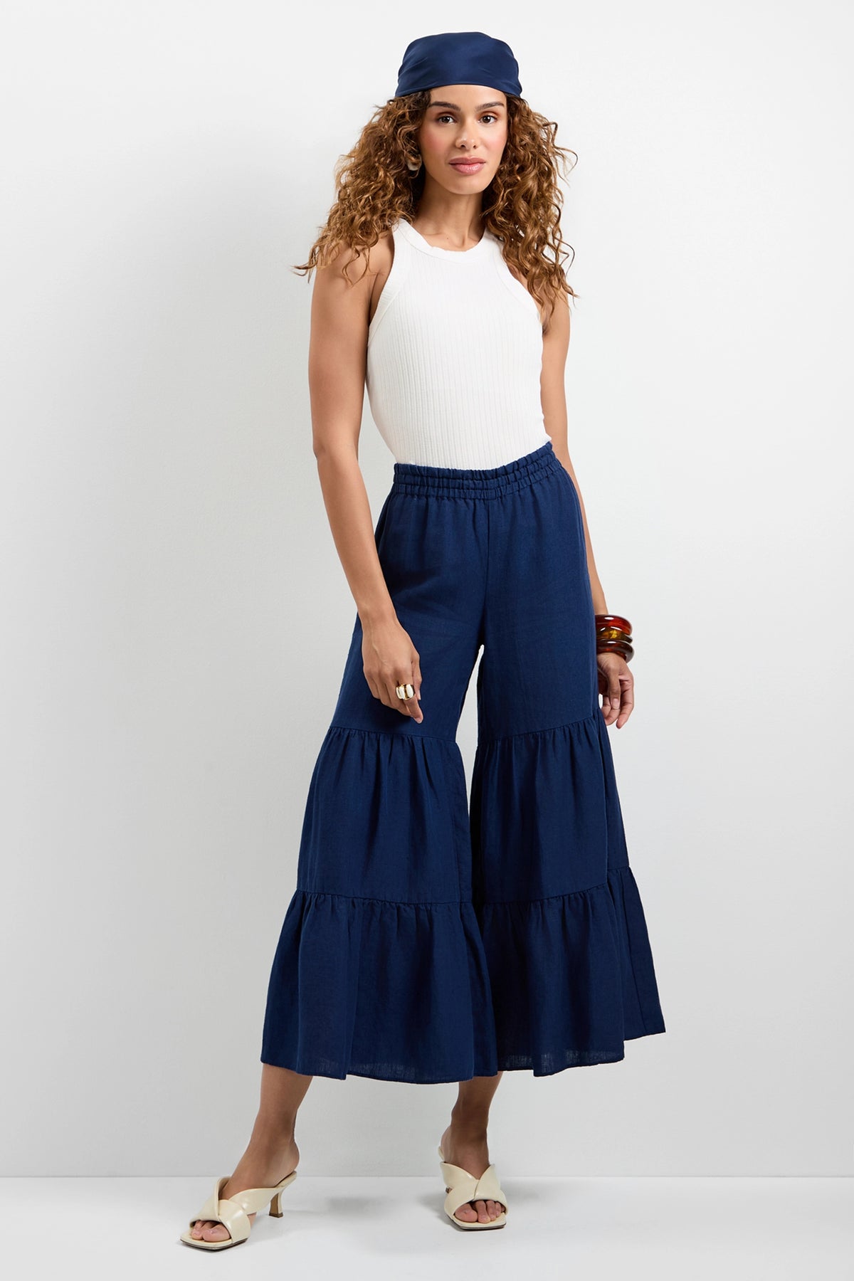 A woman with curly hair and a relaxed expression stands against a white wall, wearing the Chill Out Tiered Wide-Leg Pants, a white sleeveless top, white heeled sandals, a navy headscarf, and brown bangles.