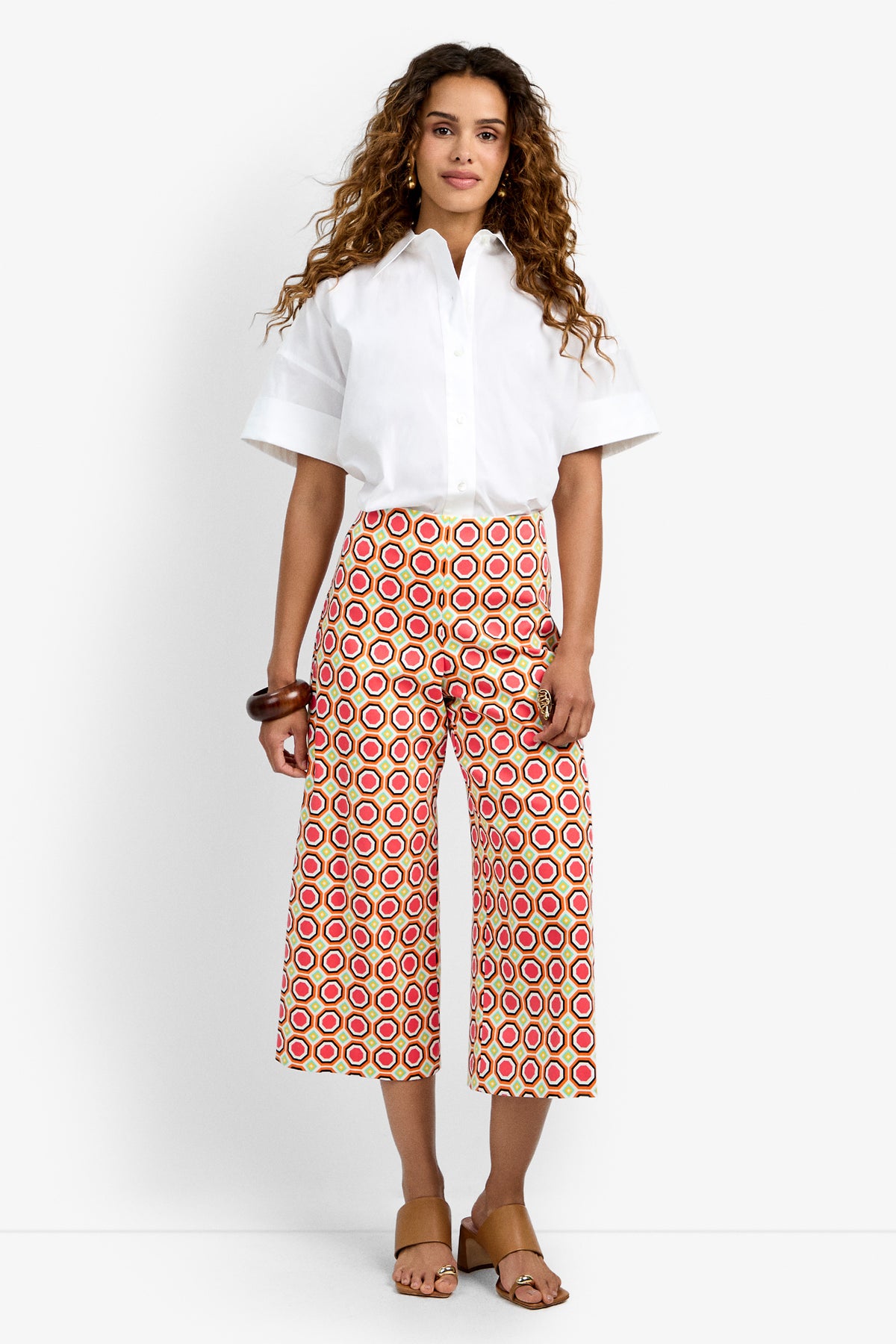 A woman with curly hair wears tan sandals, a brown bracelet, a white short-sleeve button-up shirt, and high-waisted Diona Crop Pants featuring a red, yellow, and white geometric pattern.