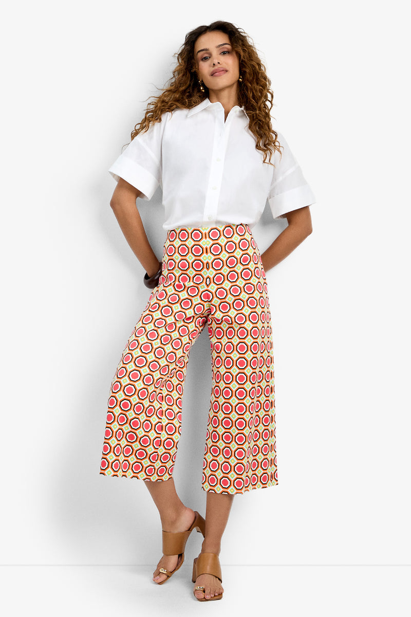 A woman with curly hair stands against a white background wearing a short-sleeve white button-up shirt, tan sandals, and the Diona Crop Pant—high-waisted, wide-leg pants featuring a bold red, yellow, and white circular pattern.