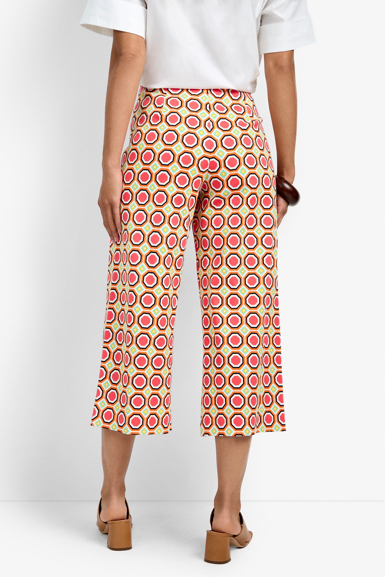A person stands facing away, wearing the Diona Crop Pant featuring a red, yellow, and white hexagon pattern. They pair it with a white short-sleeve shirt and tan heeled sandals.
