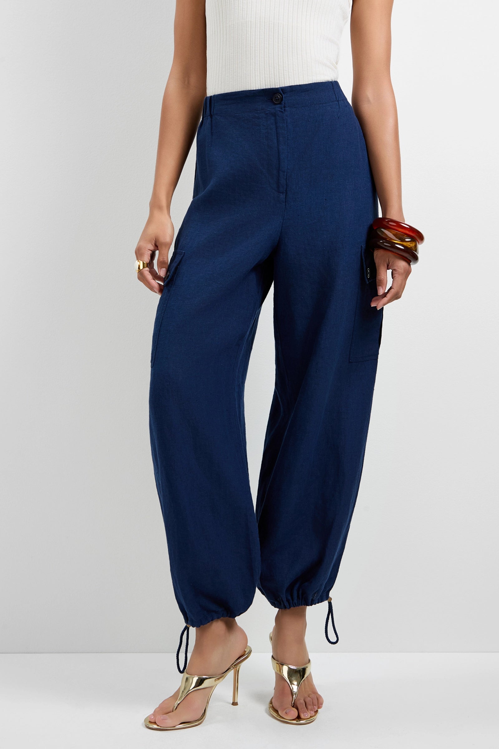 A person wearing Alico Cargo Pants in dark blue, paired with a white sleeveless top, gold high-heeled sandals, and chunky bracelets stands against a plain light background.